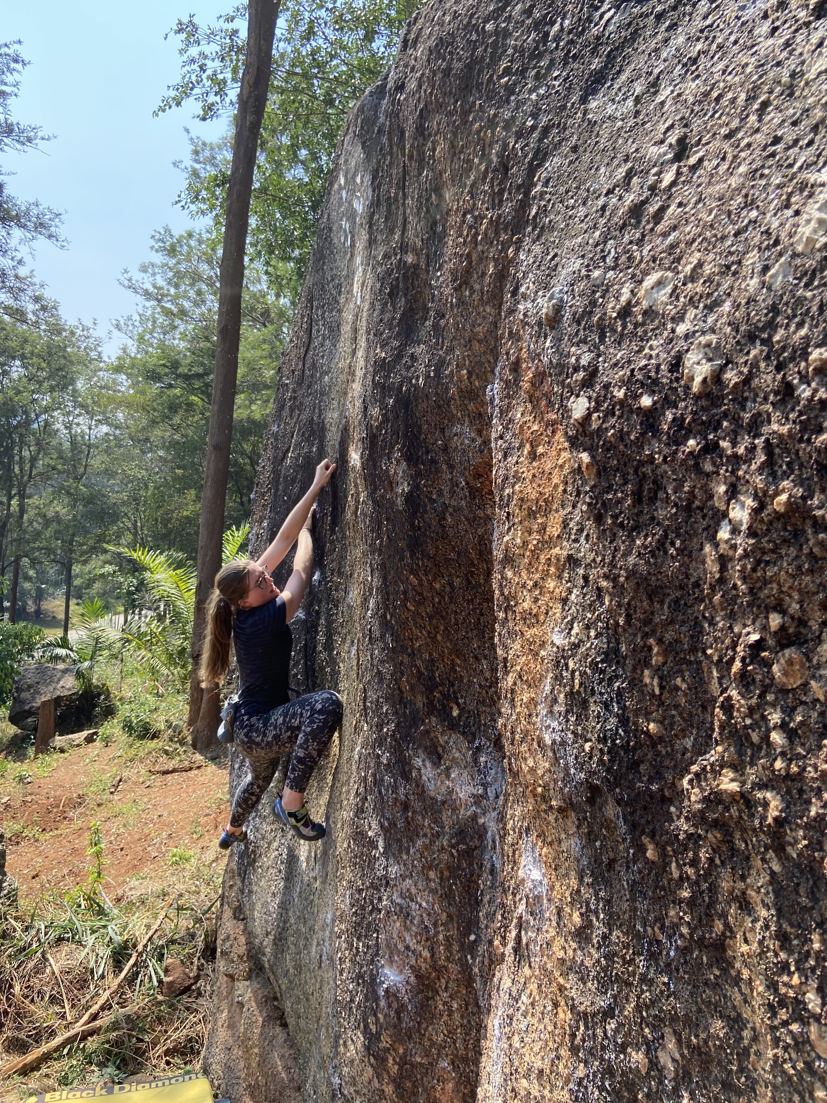 Climber crimping above a bouldering pad on a granite boulder set within a coffee plantation.