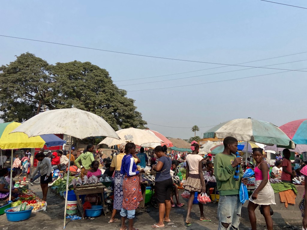 Busy and bustling market with people selling and buying different fruit and vegetables under parasols.