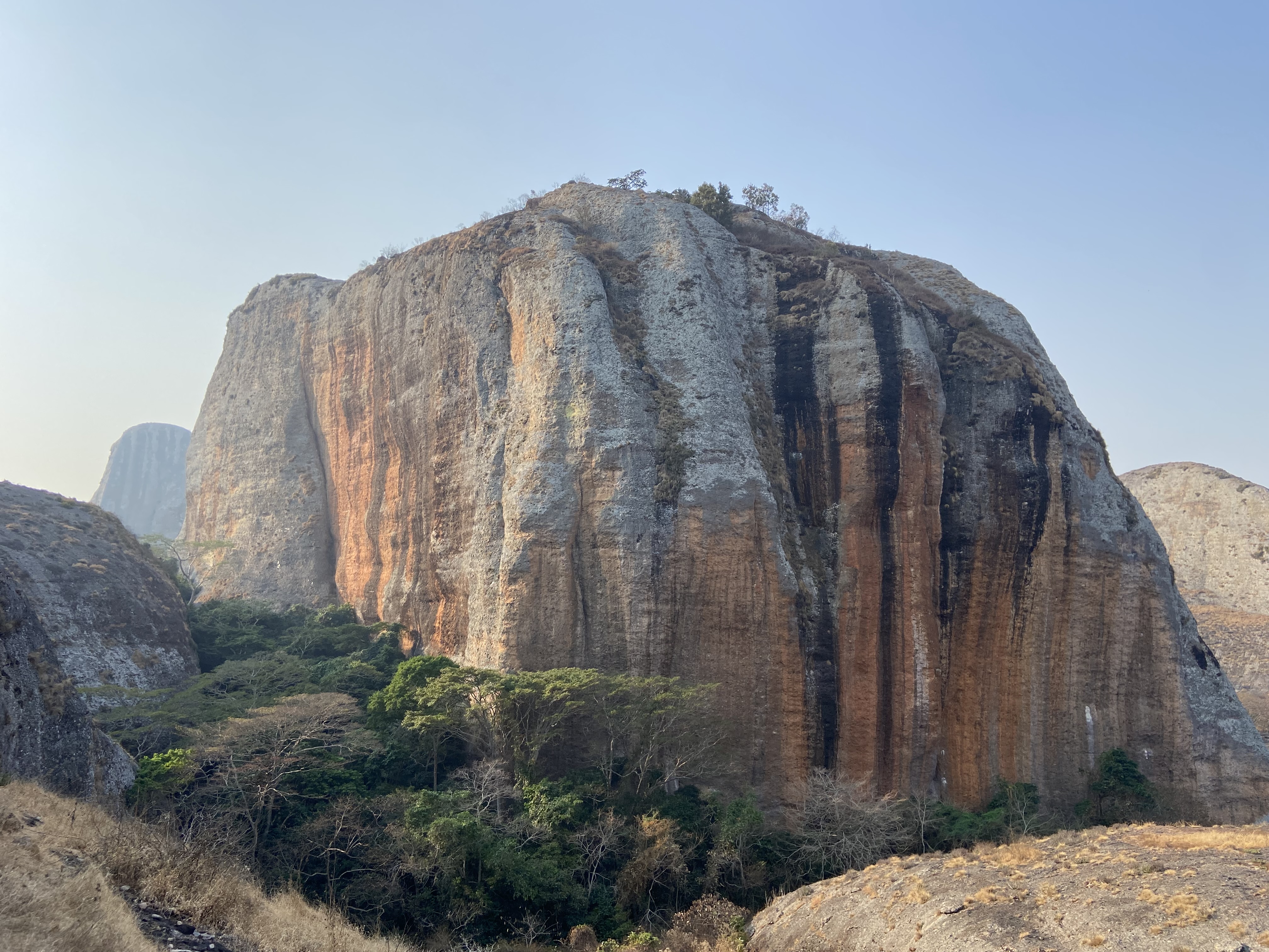 Large prominant cliff with grey and orange rock with black streaks running down the grooves in the rock face.