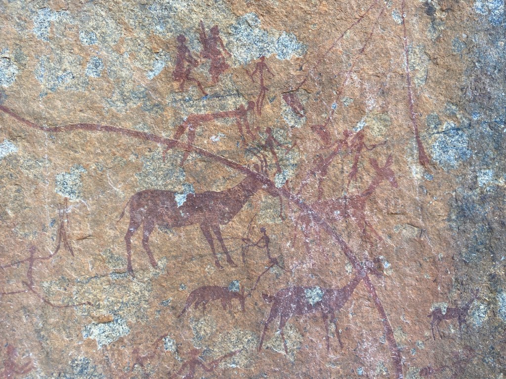 Red cave painting depicting people with spears and different animals such as antelopes and an aardvark. 