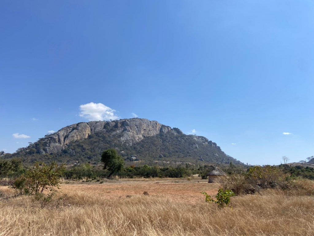 Large granite cliffs rising out of the savanna with a small rondavel in the foreground. 
