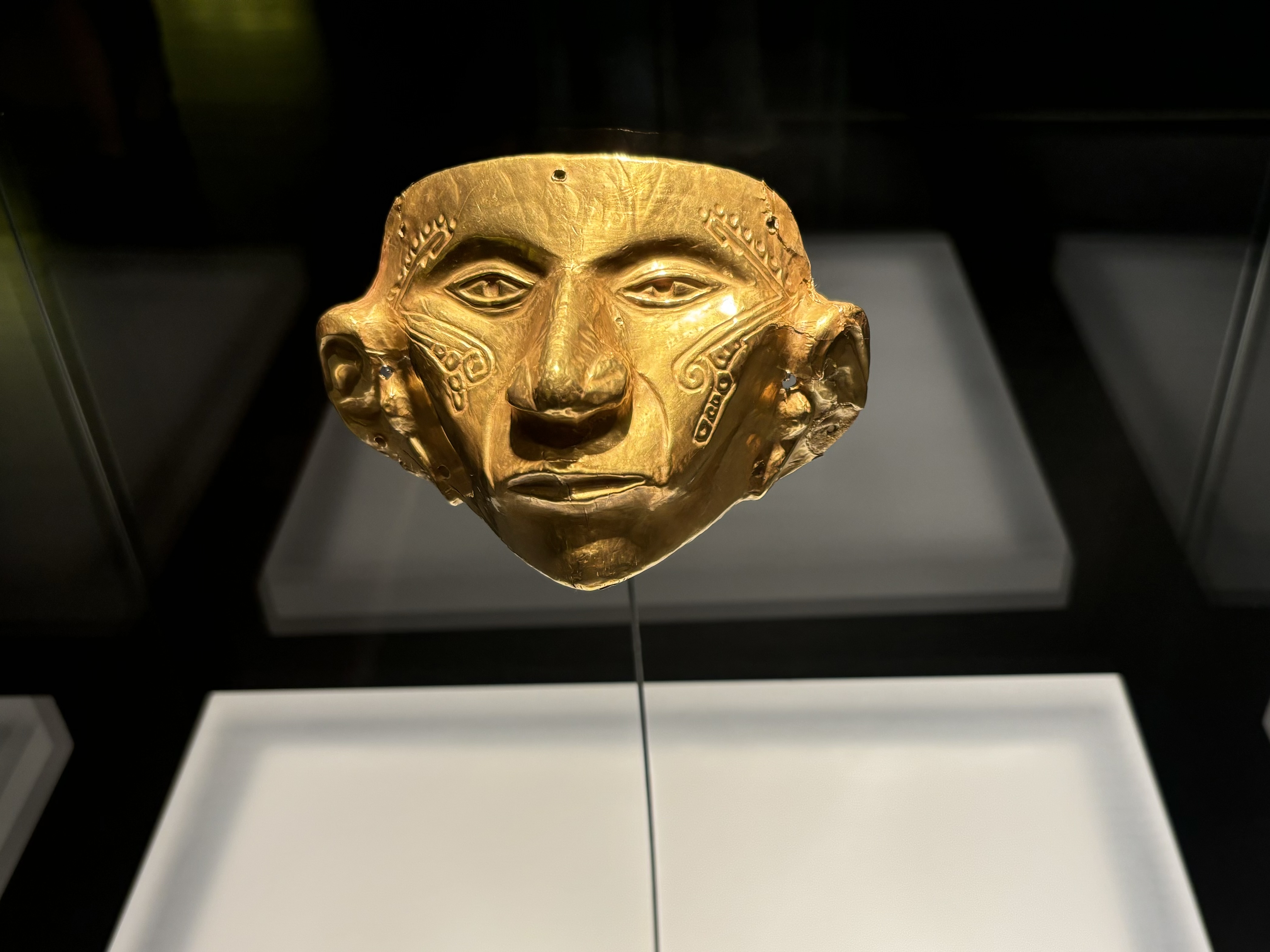 Mask of a man made from gold with ornate patterns across his temples and cheeks.