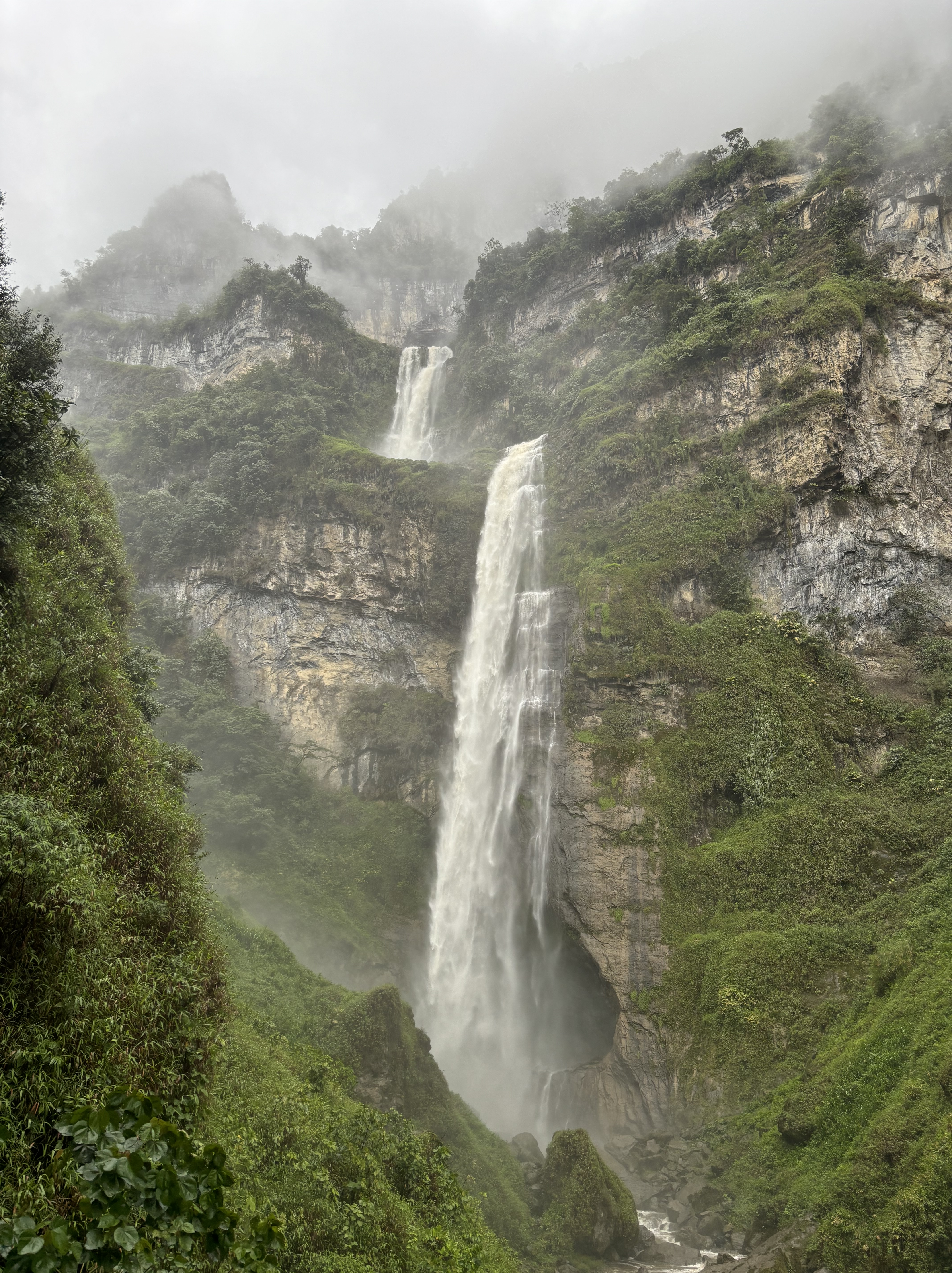 Moody and misty scene with limestone cliff covered in lush green vegetation with a two tiered fast flowing waterfall.