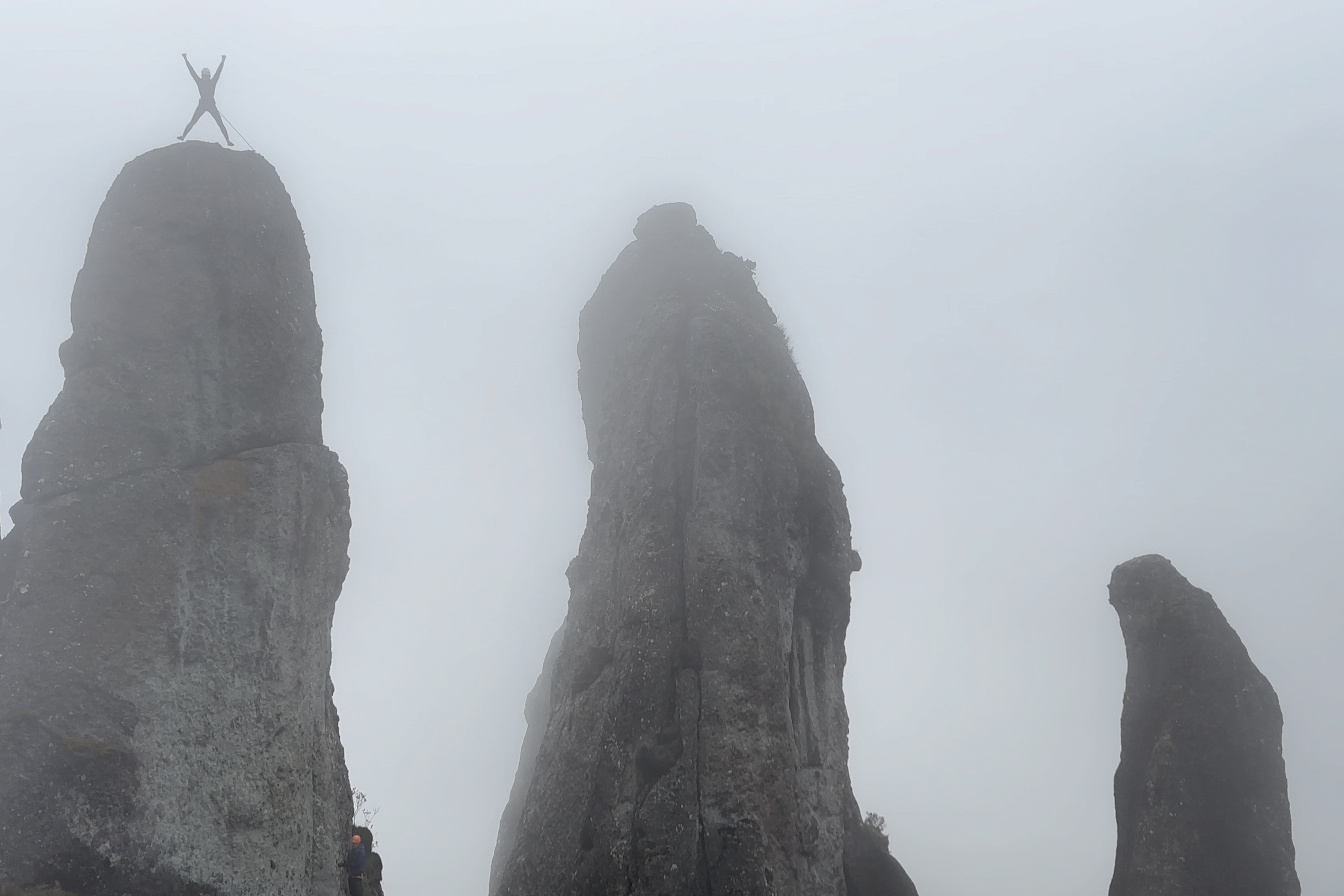 Climber jumping at the top of one of three finger like pillars set within thick fog. 