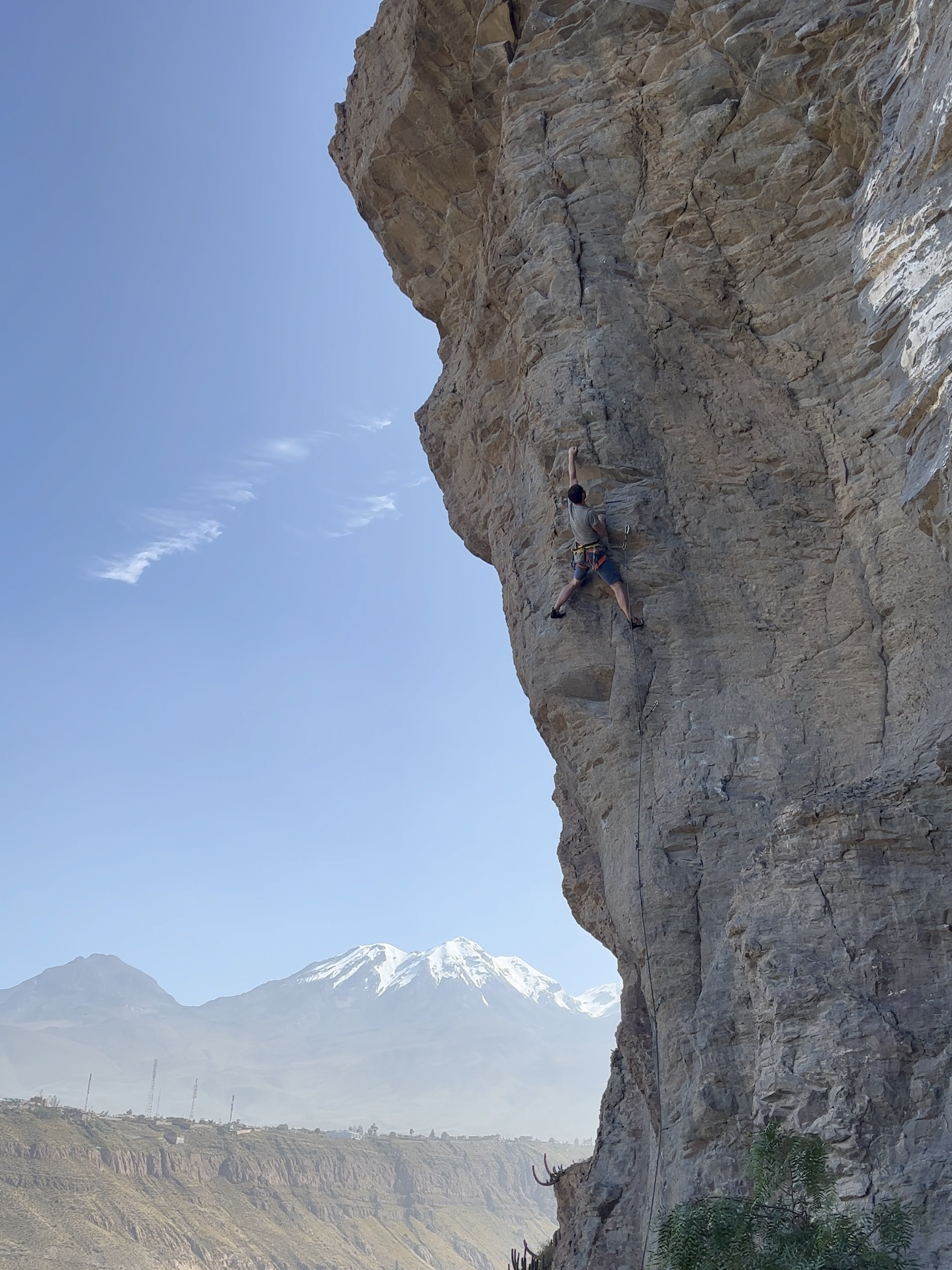 Climber reaching high on steep grey rock with snowy mountain peaks in the background. 