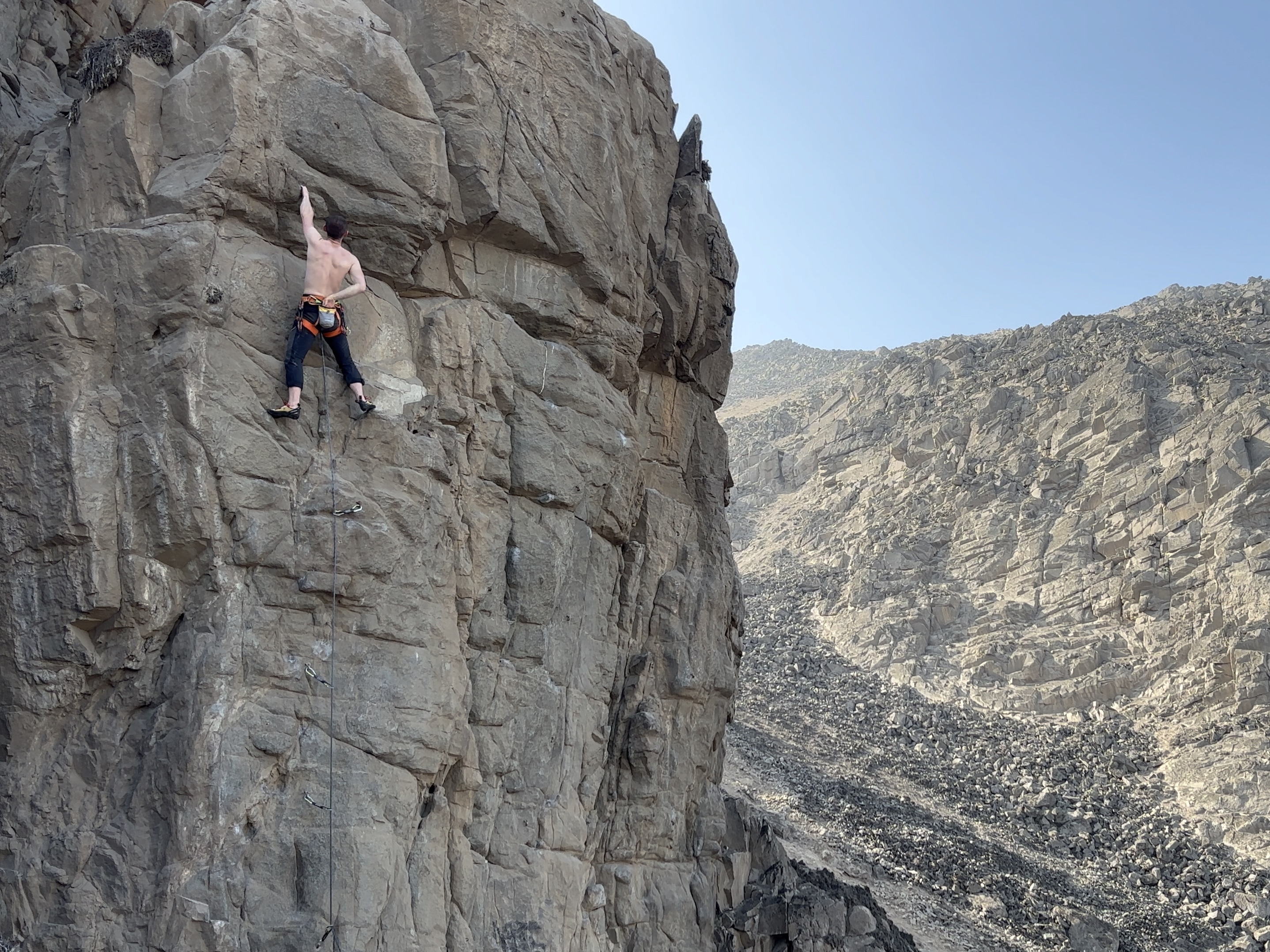 Climber hanging on one hand to chalk up on the vertical grey rock set within the dry, desert environment. 