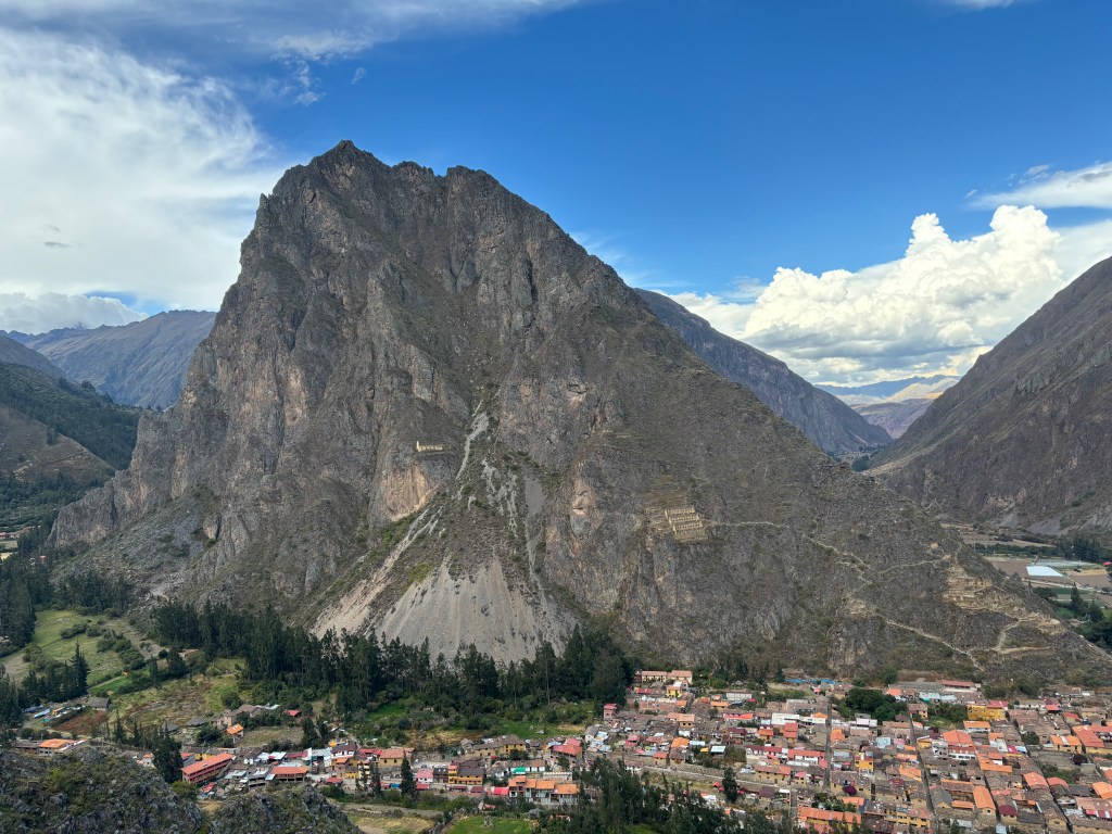 Ancient stone building set upon a steep sided mountain above a town of orange roof tiled houses. 