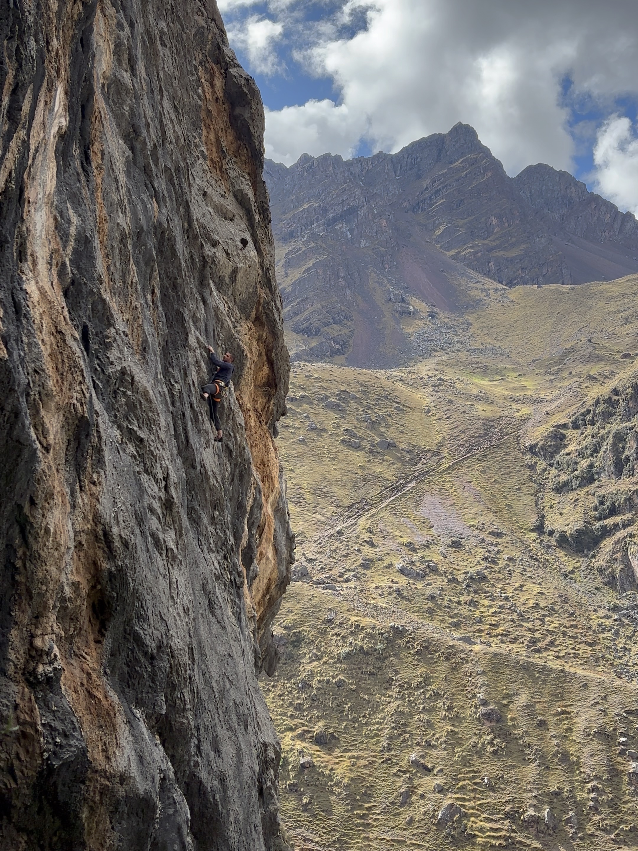 Climber reaching for the next handhold on a vertical limestone wall streaked with grey and orange, with jagged mountain peaks forming a narrow valley in the background.