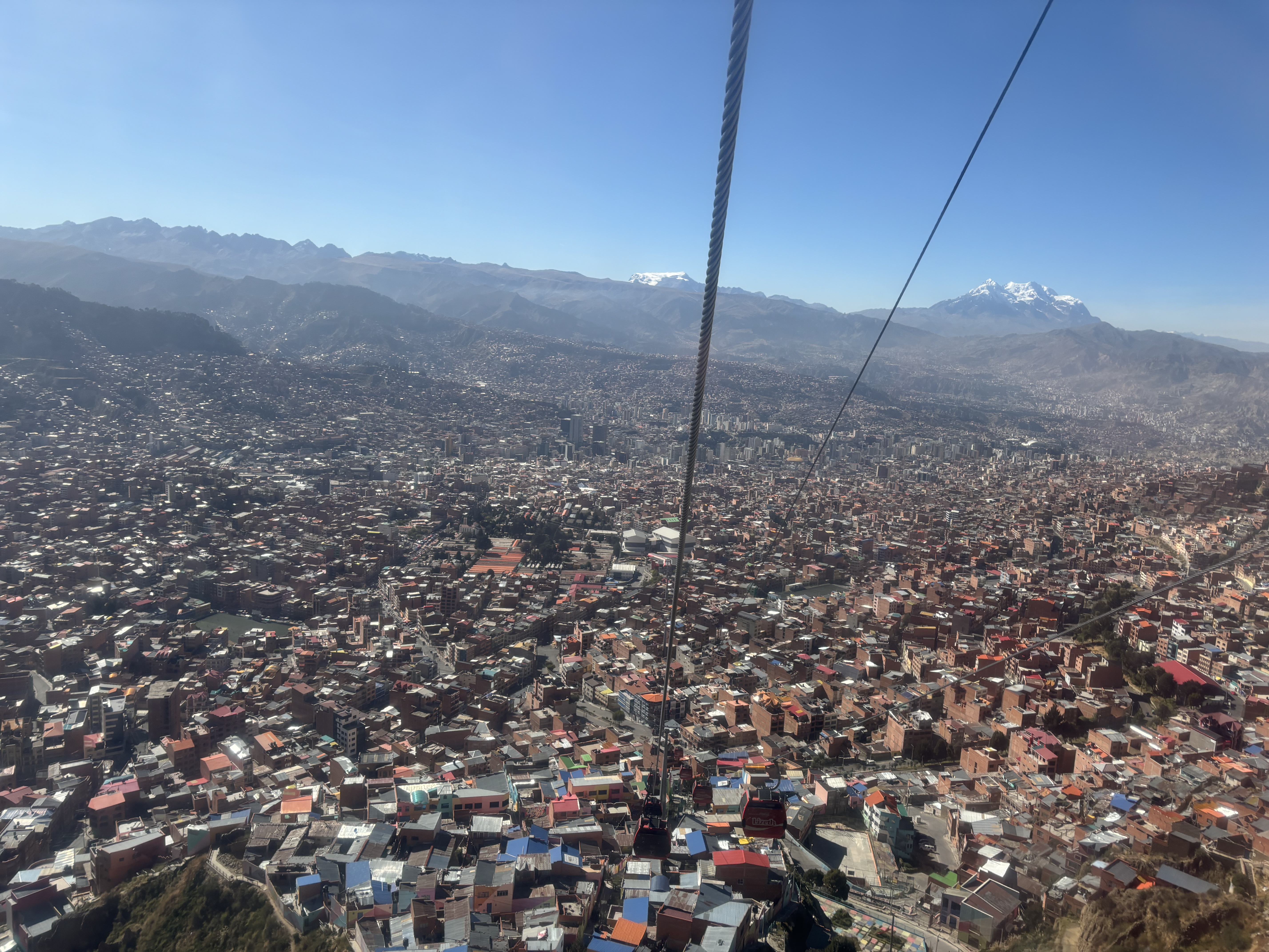 Cable cars running above tightly packed buildings in the city below, with high-altitude snowy peaks visible in the distance.