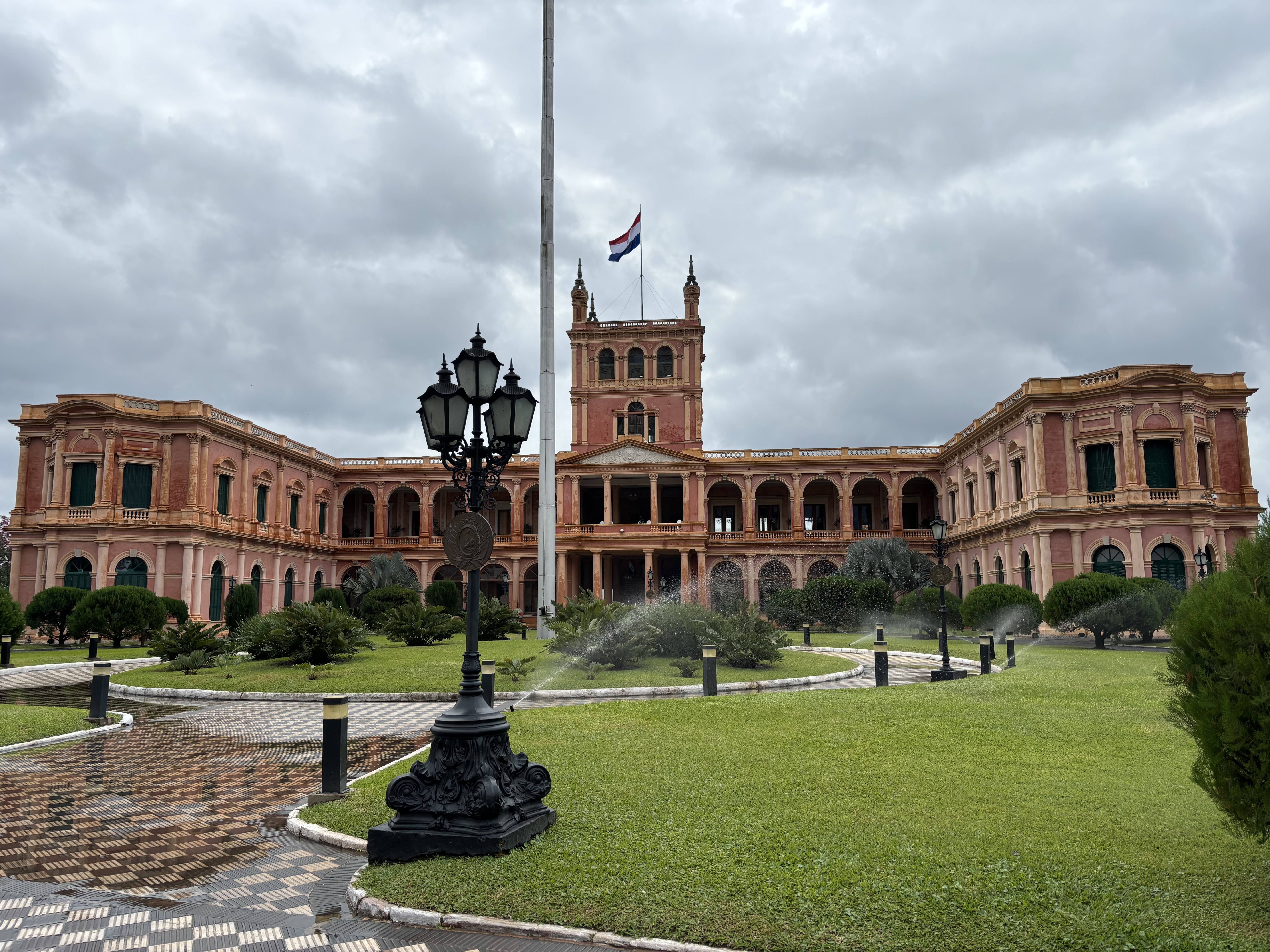A grand pink palace with a huge Paraguayan flag on top in front of a  green manicured lawn with ornamental trees.