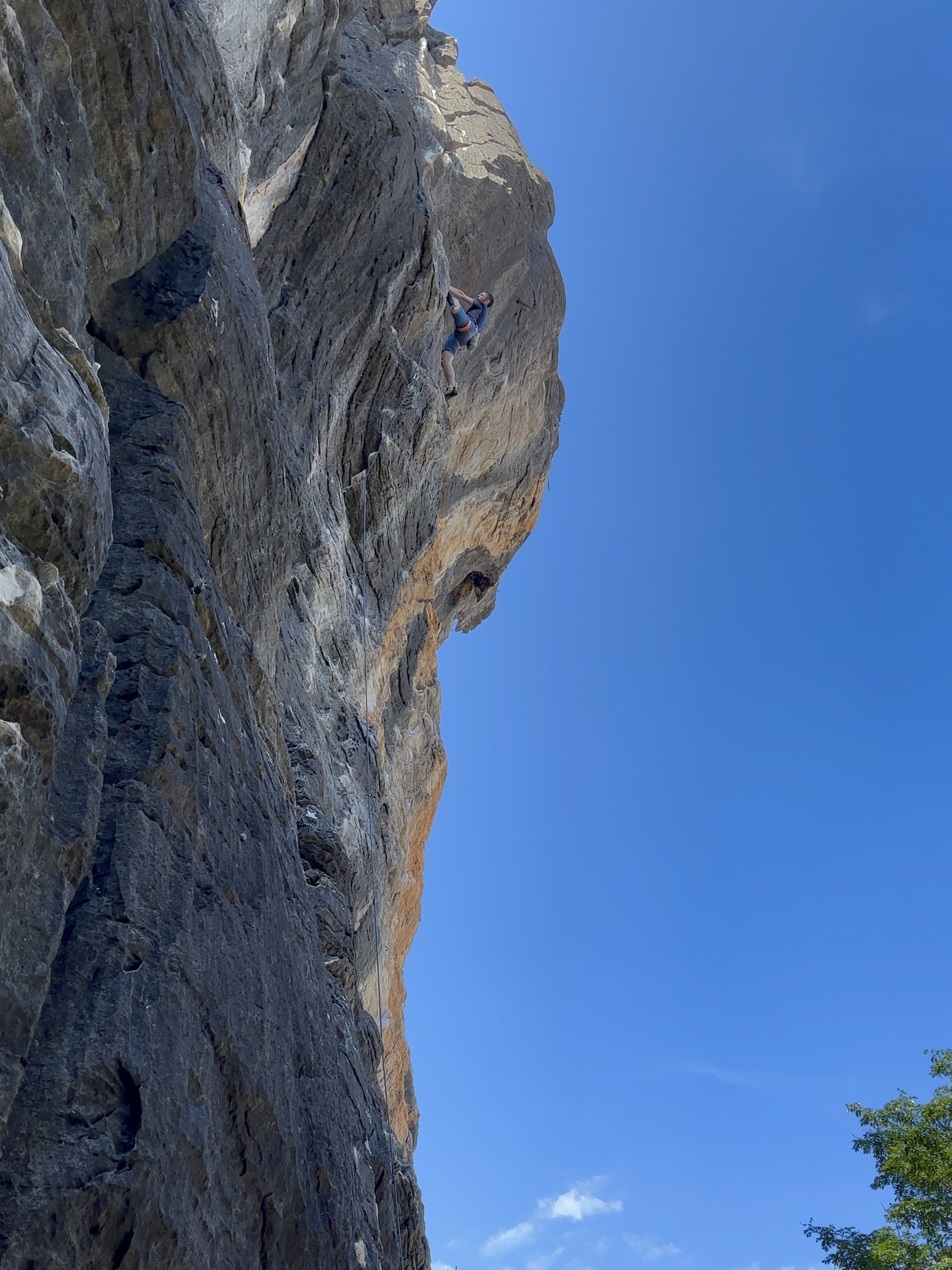 Climber on an overhanging, limestone/marble wall with grey and yellow streaks, set against a bright blue sky in the background. 