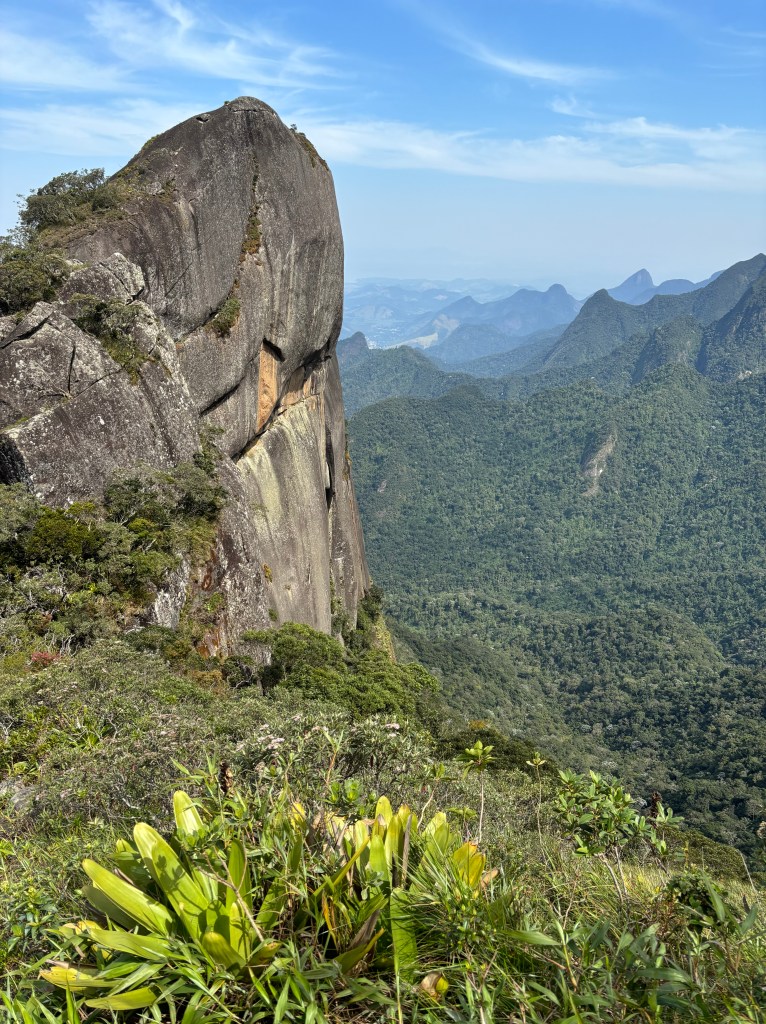 Small rock pinnacle on the top of a tall mountain with other forested mountain peaks in the background.
