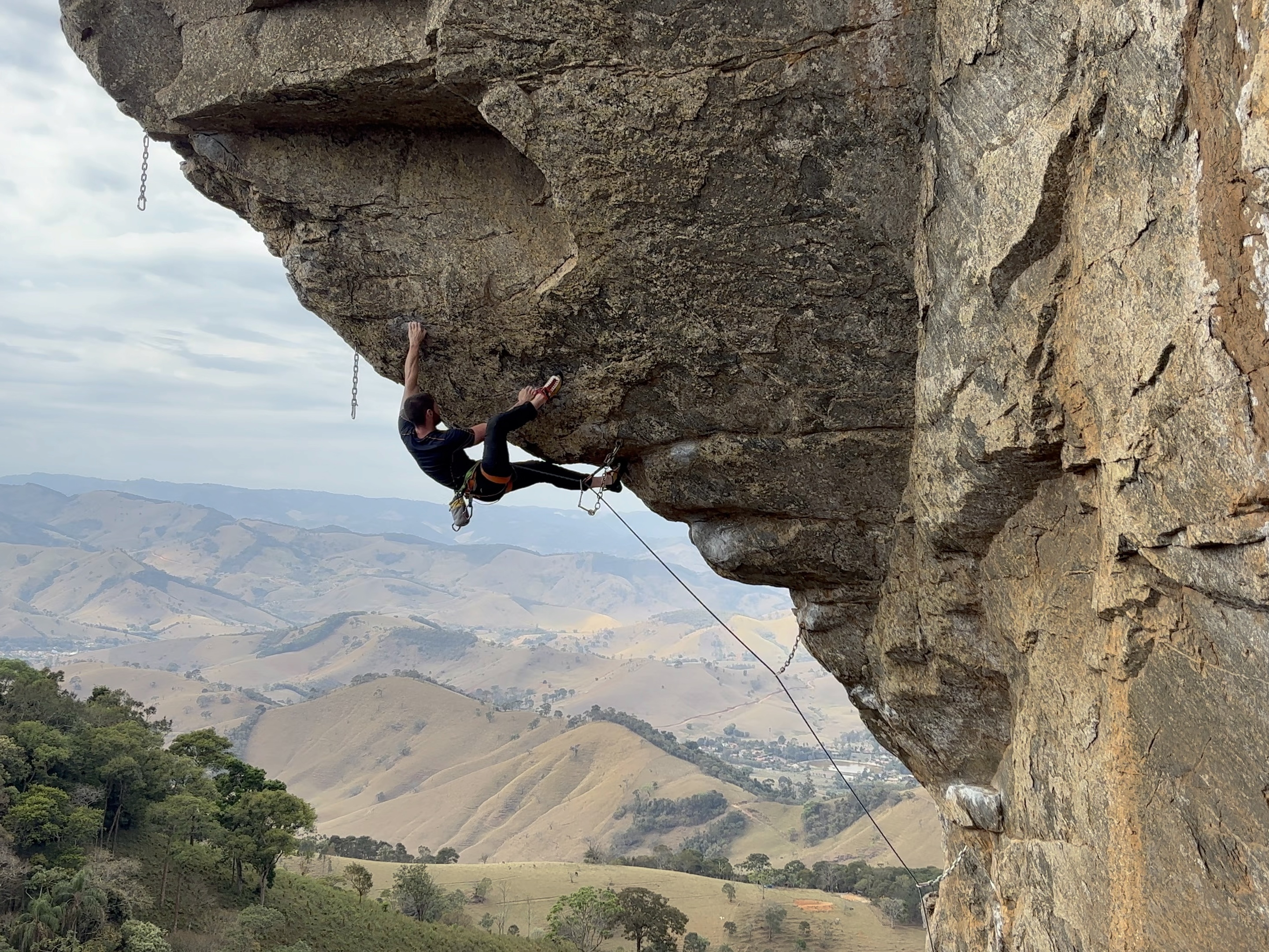 Climber heel hooking on a steep, overhanging arete overlooking rounded hills and forest below.