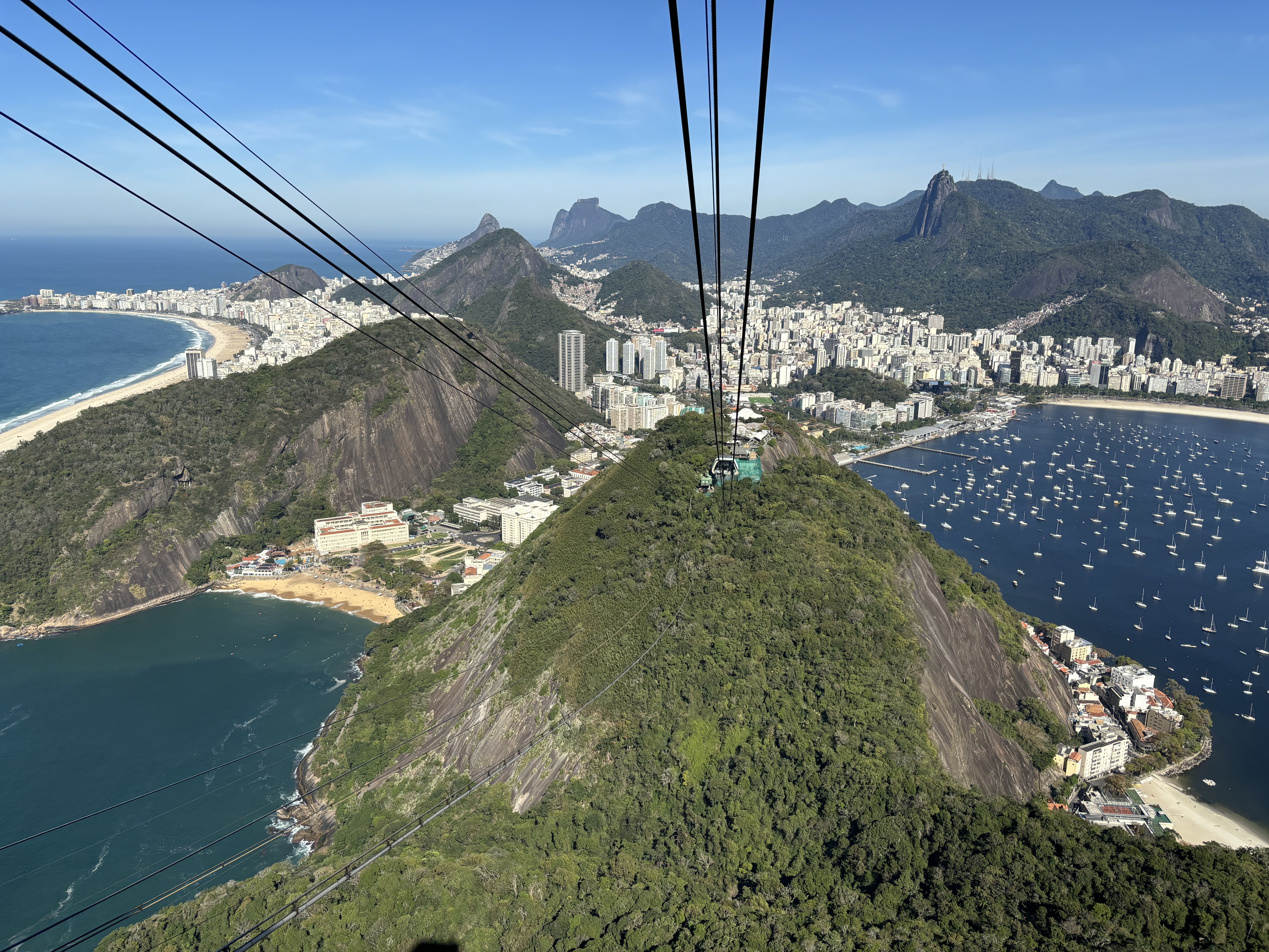 Steep cable car lines running between two granite domes overlooking the city of Rio de Janeiro, with high-rises, granite formations and sandy beaches below.