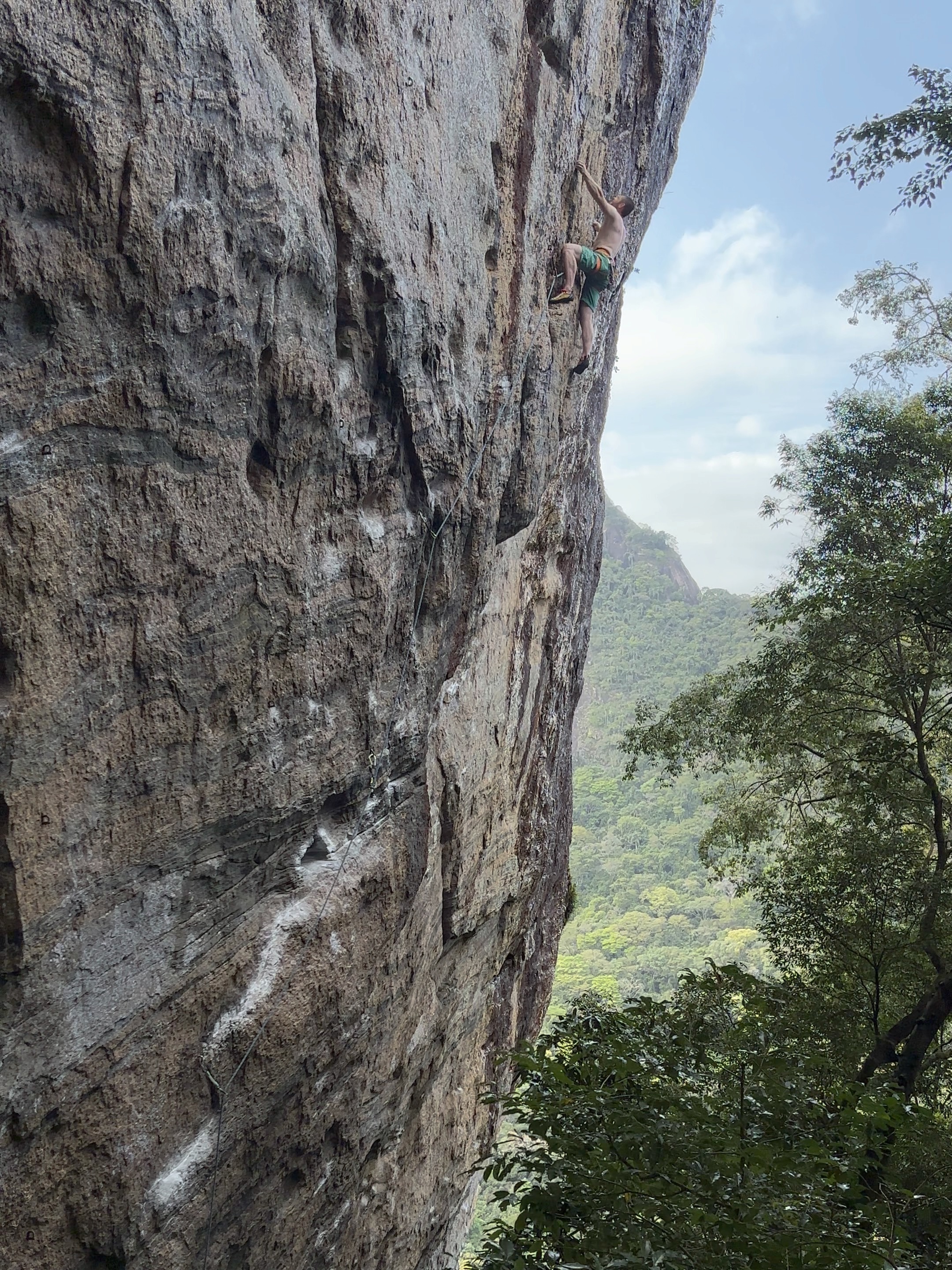 Climber in green shorts on an overhanging, gneiss sport climb with a green mountainside in the background.