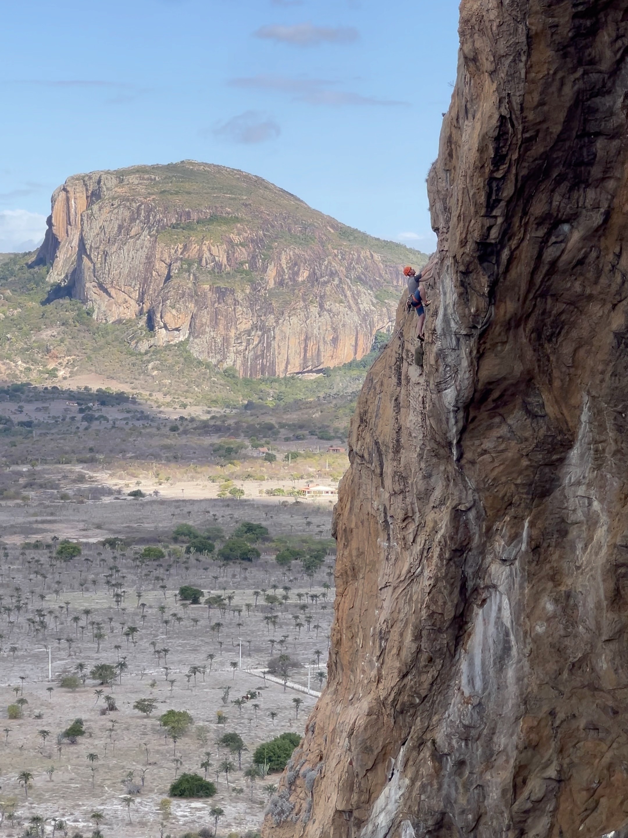 Climber in an orange helmet climbing on the lip of a gneiss cave with dramatic cliffs in the background set in a flat semi arid landscape dotted with green trees.