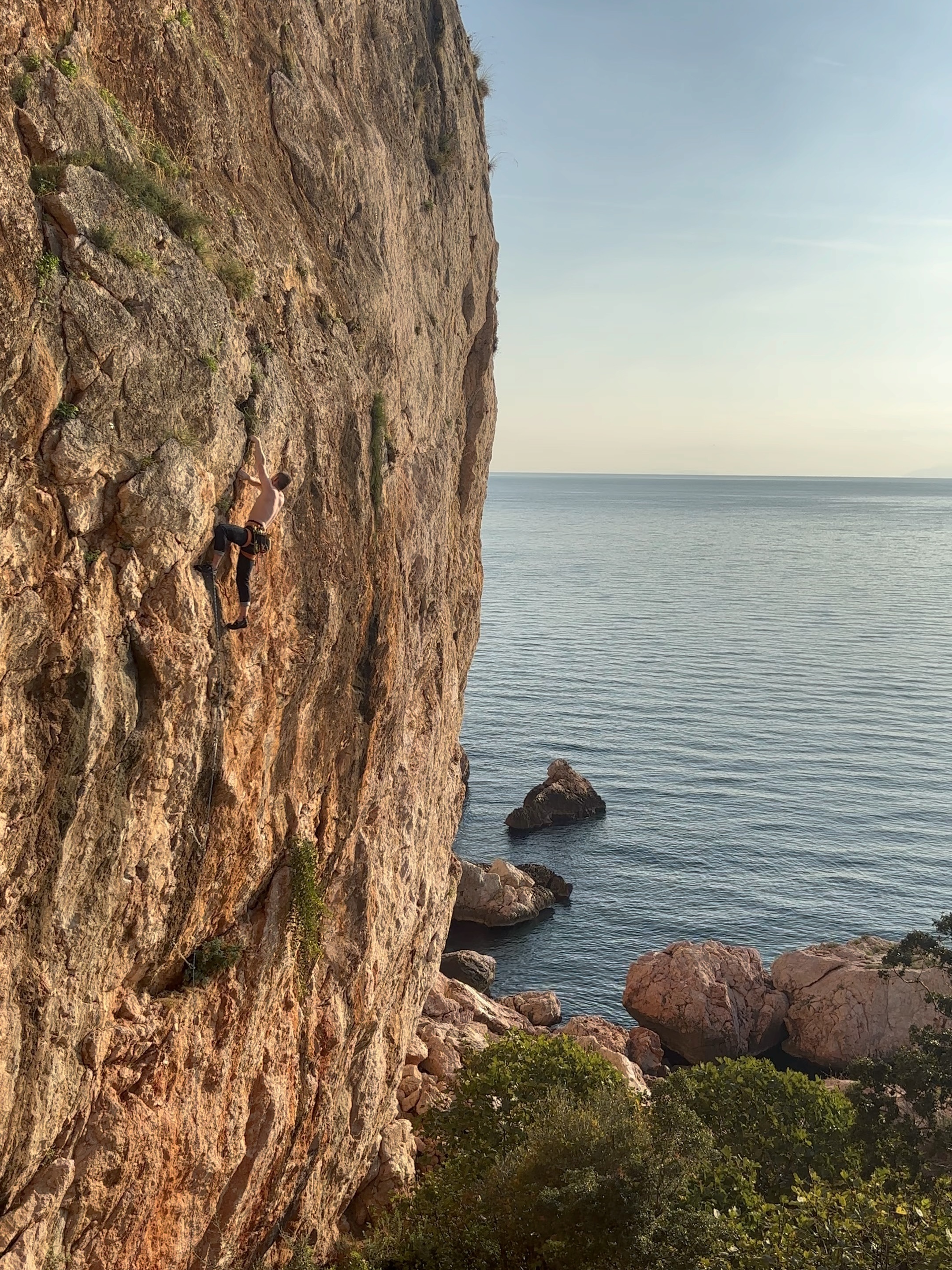 Climber laybacking a flake feature on an orange limestone cliff, directly above the sea.