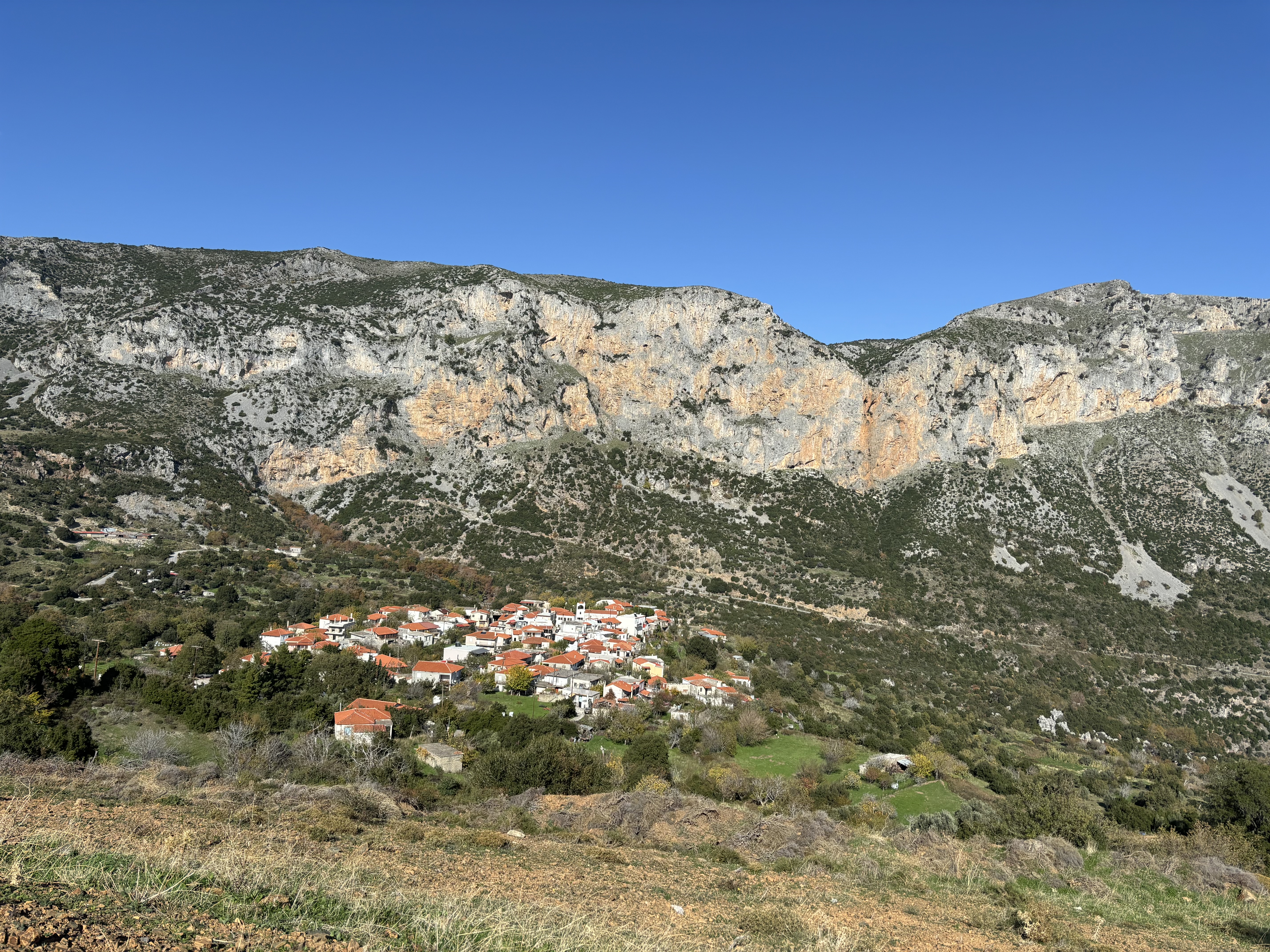 A large band of limestone cliffs rising above a small greek village of white-washed houses with terracotta roofs.