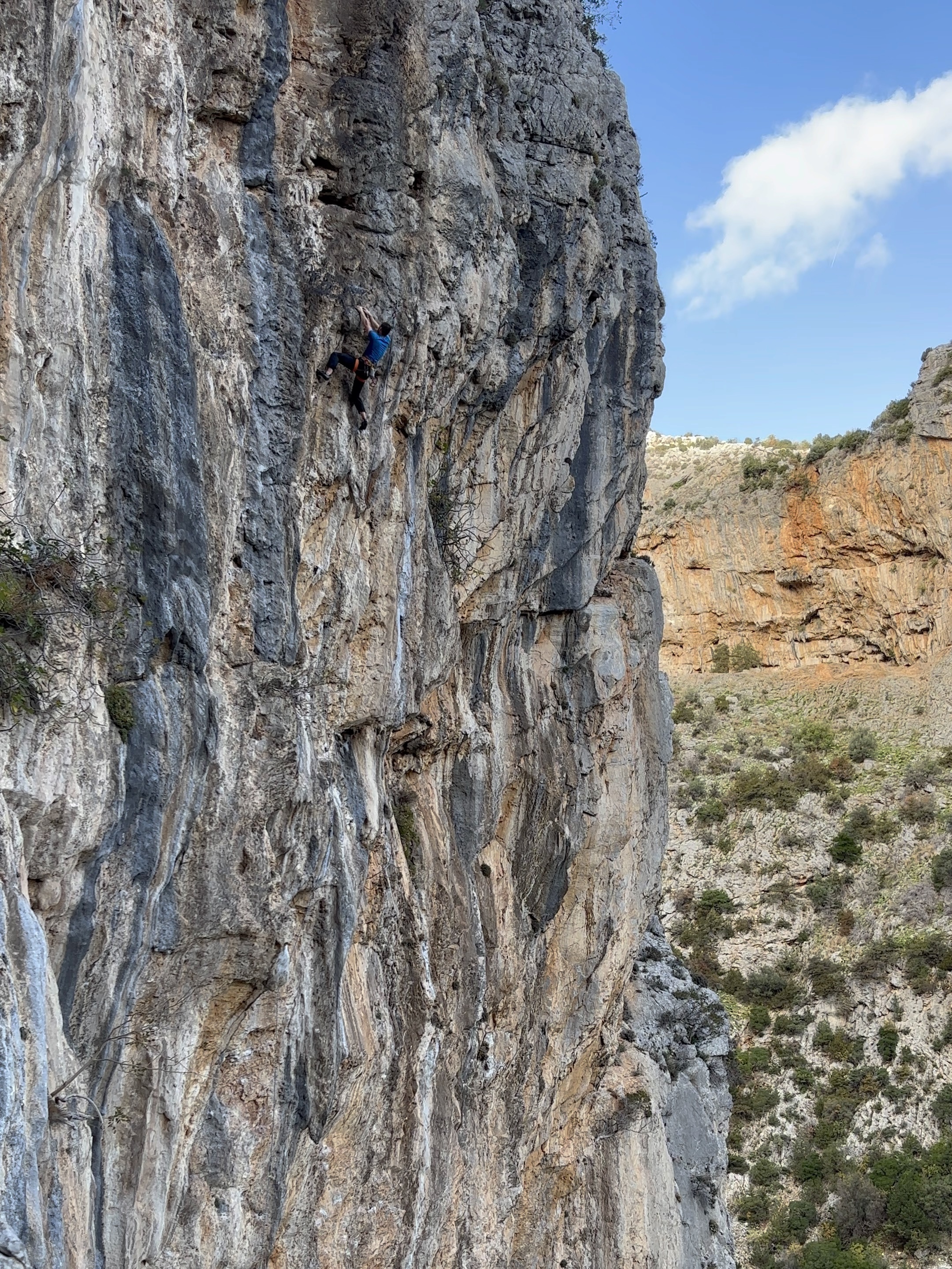 Climber using a high left foot on a tall grey wall, with large orange cliffs in the background.