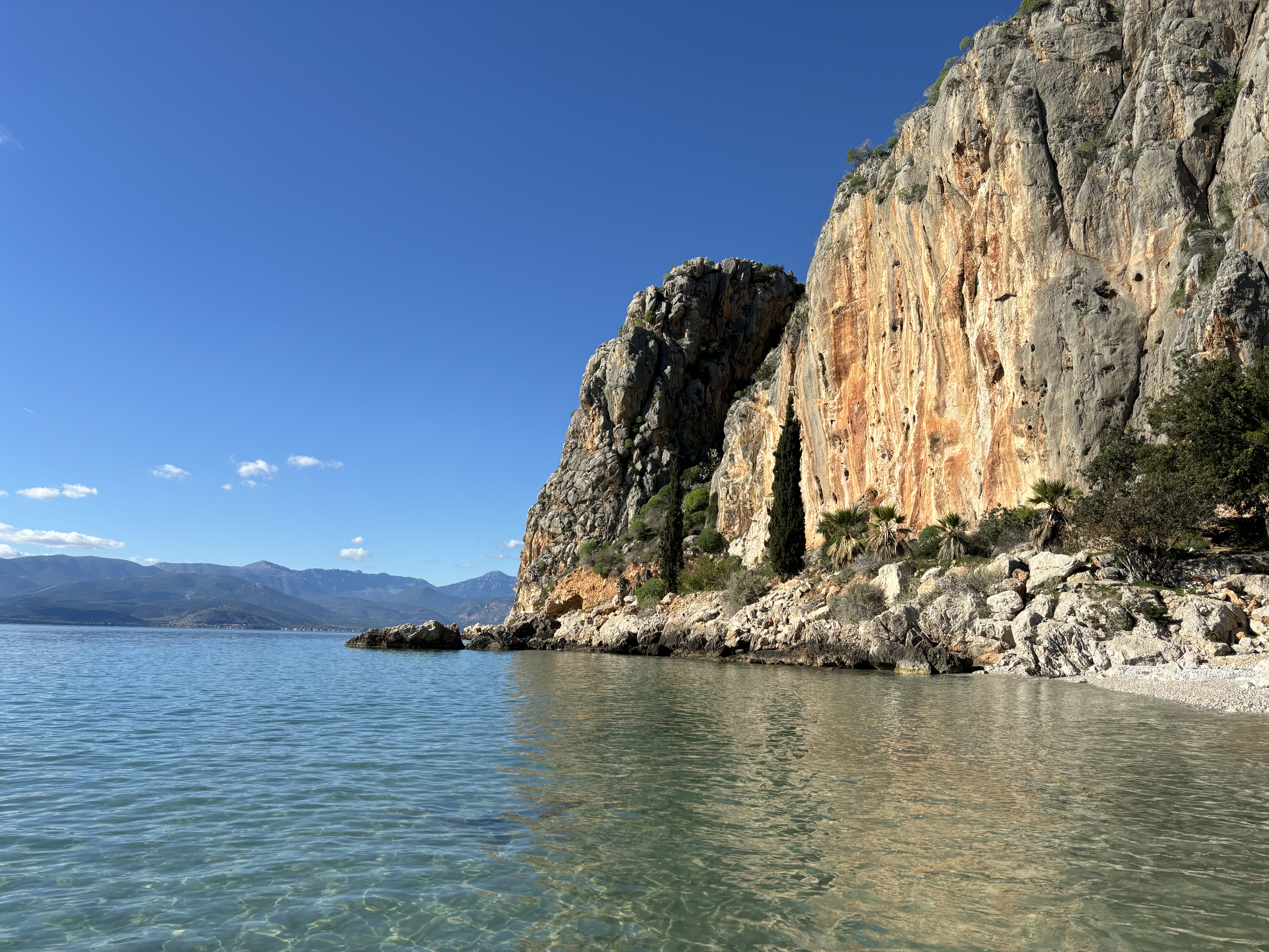 Orange sea cliff set on the edge of the crystal clear water, with mountains in the background.
