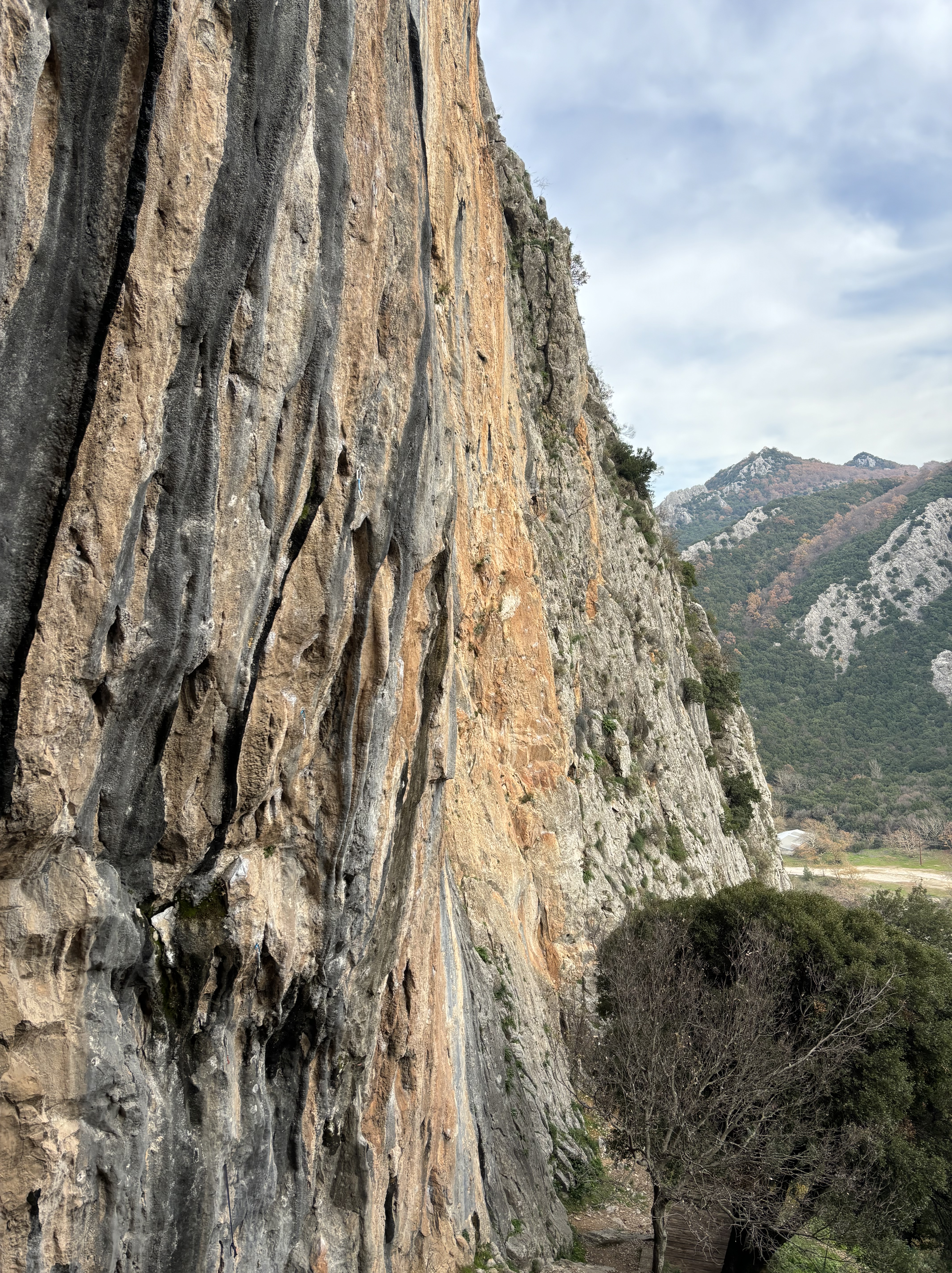 Tall orange limestone wall covered in dark grey and black tufas, with mountains in the background.