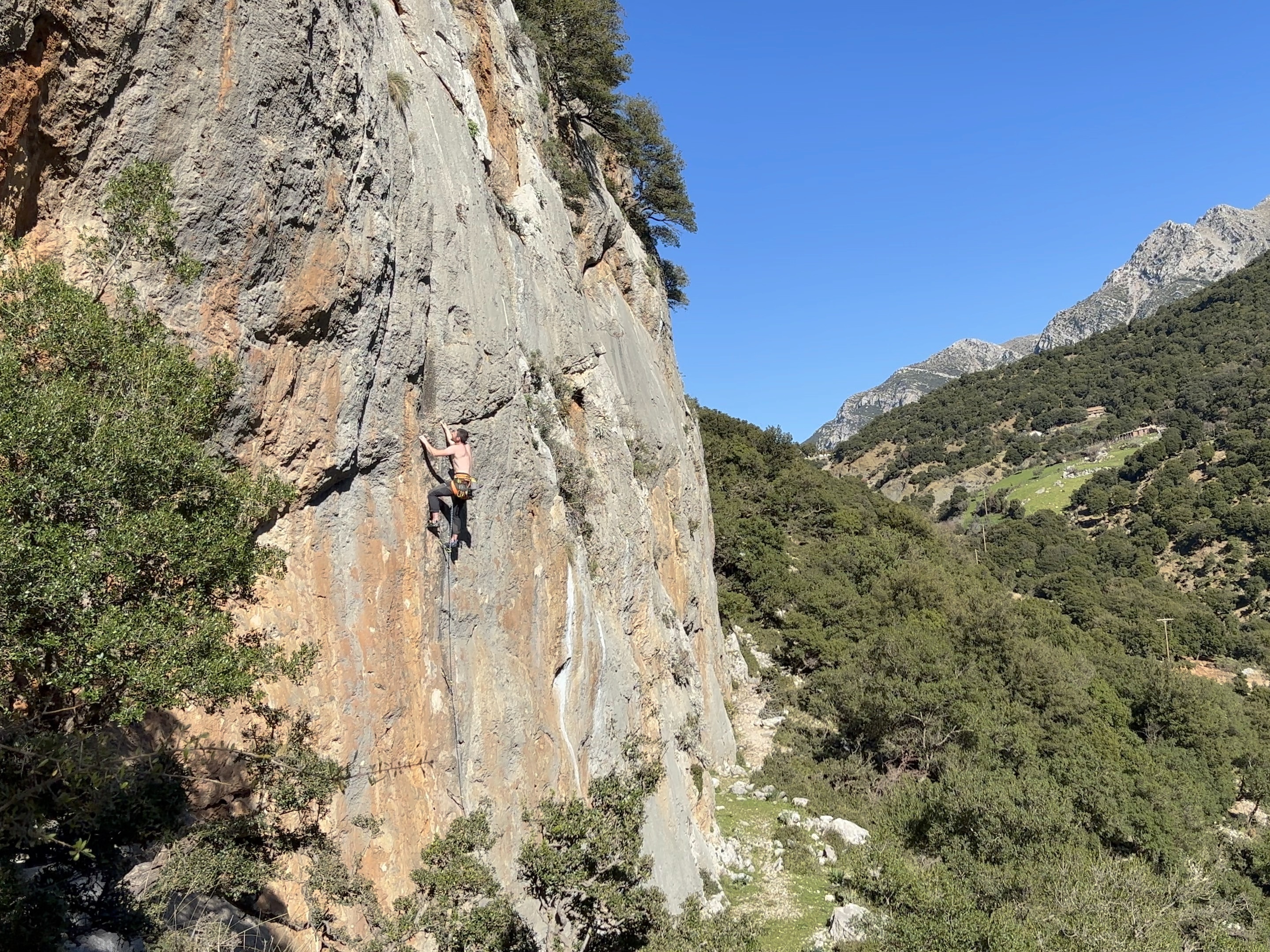 Climber on a grey and orange limestone wall set in a lush, green, mountainous area.