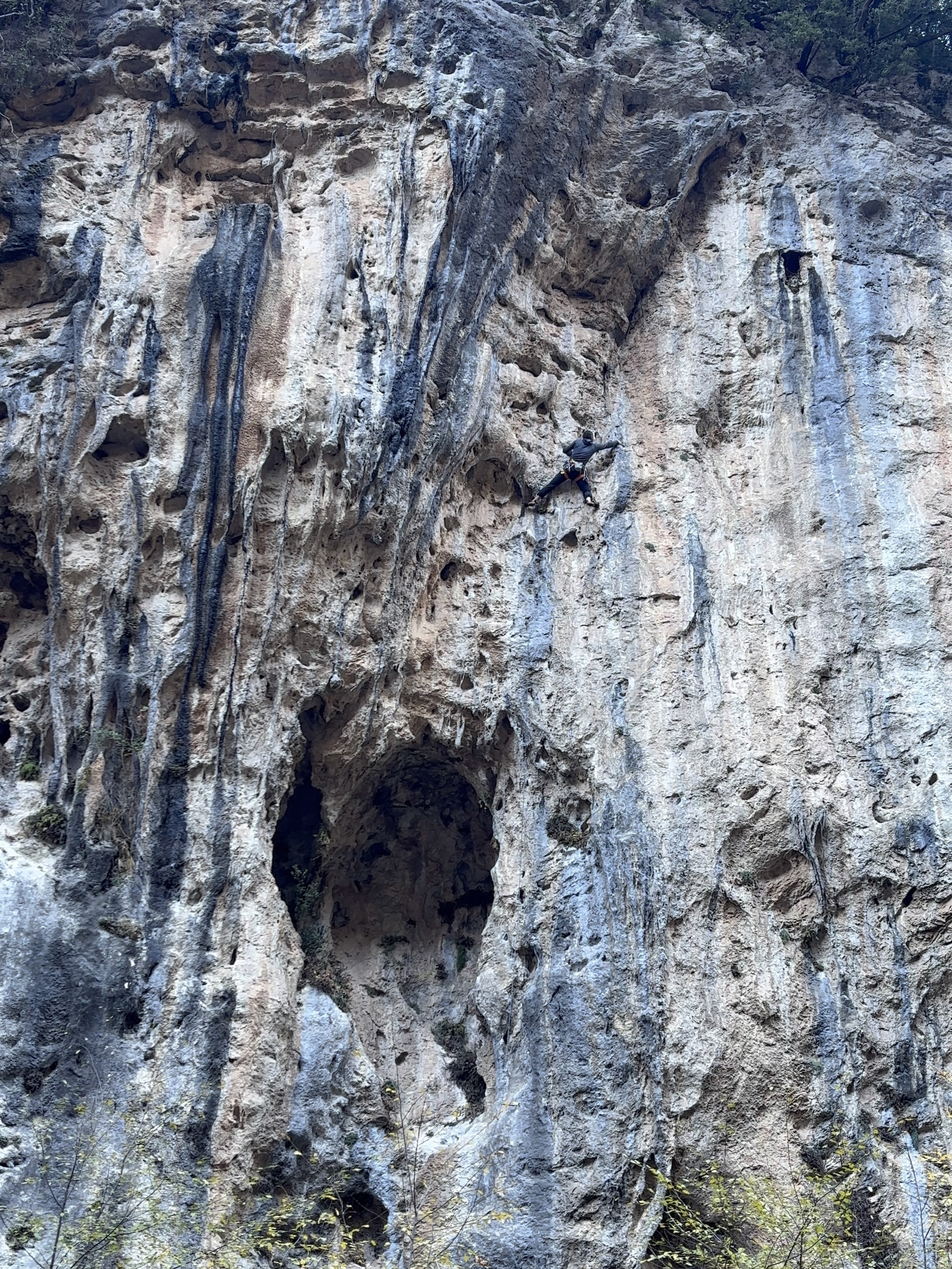 Climber in a grey fleece reaching out right above a small cave set within a vertical wall.