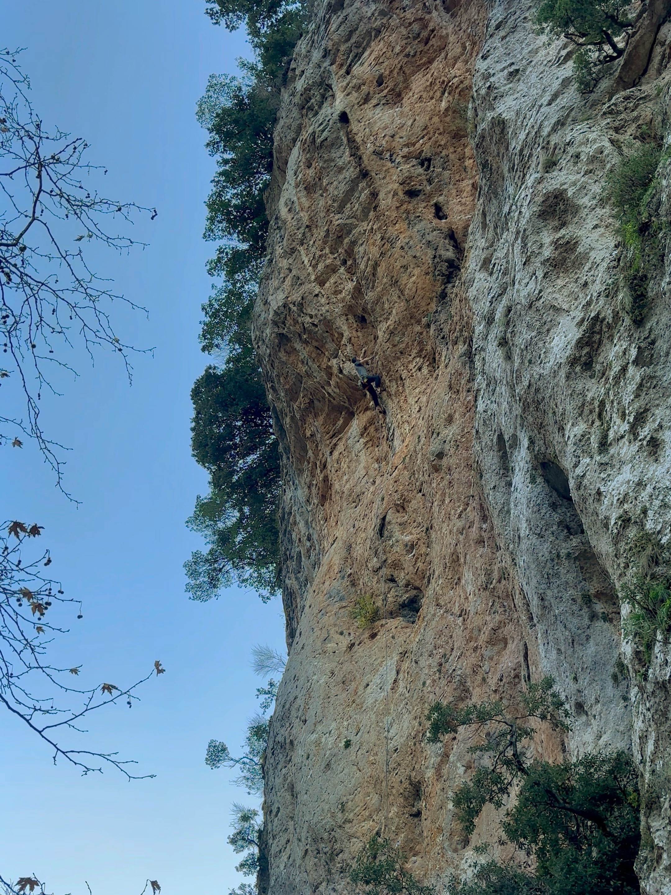 Climber tackling an overhanging section of rock at the top of a tall orange limestone cliff.