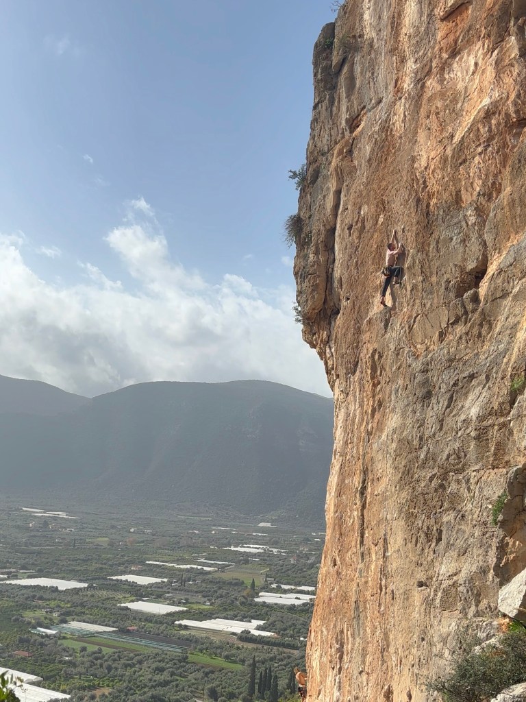 Climber on a vertical limestone wall above agricultural fields, with a steep sided mountain in the background.