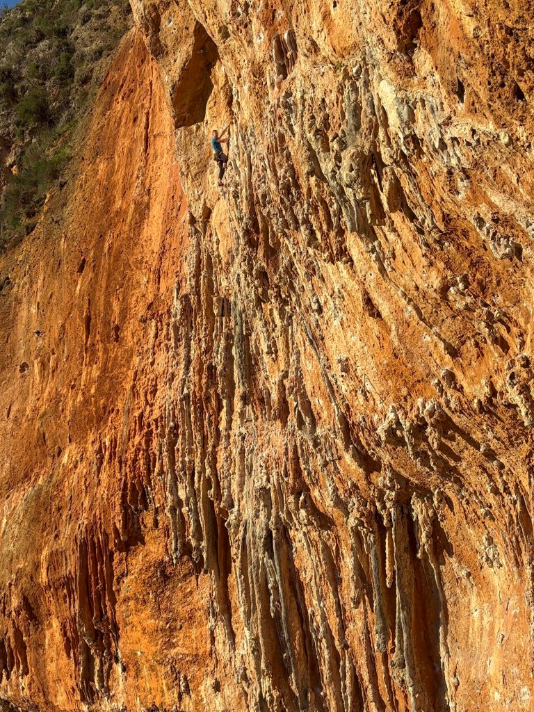 Climber lost within a sea of tufas on a steep orange limestone cliff.
