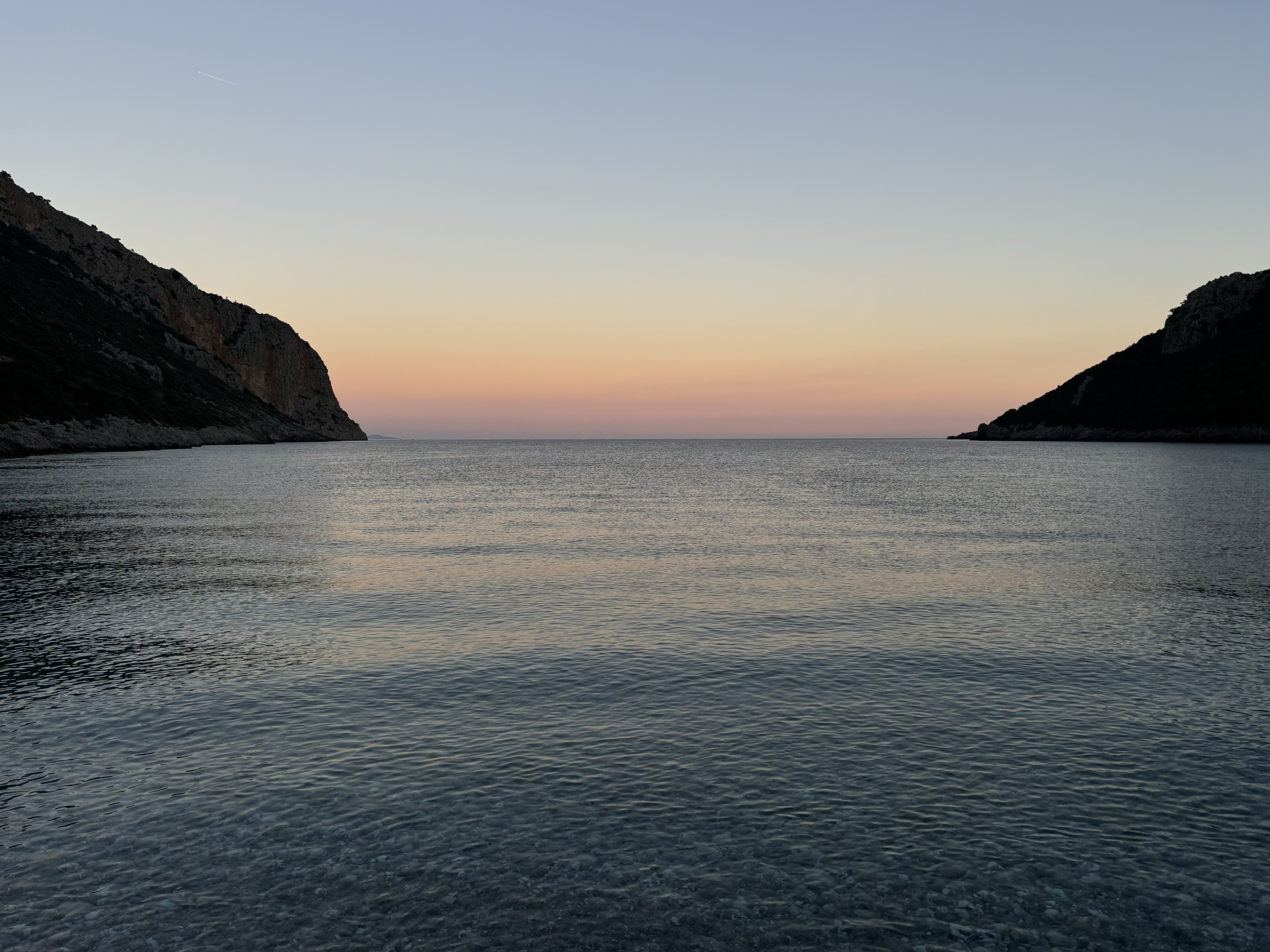 Pale pink sunset over calm ocean water, with cliffs forming a sheltered bay.
