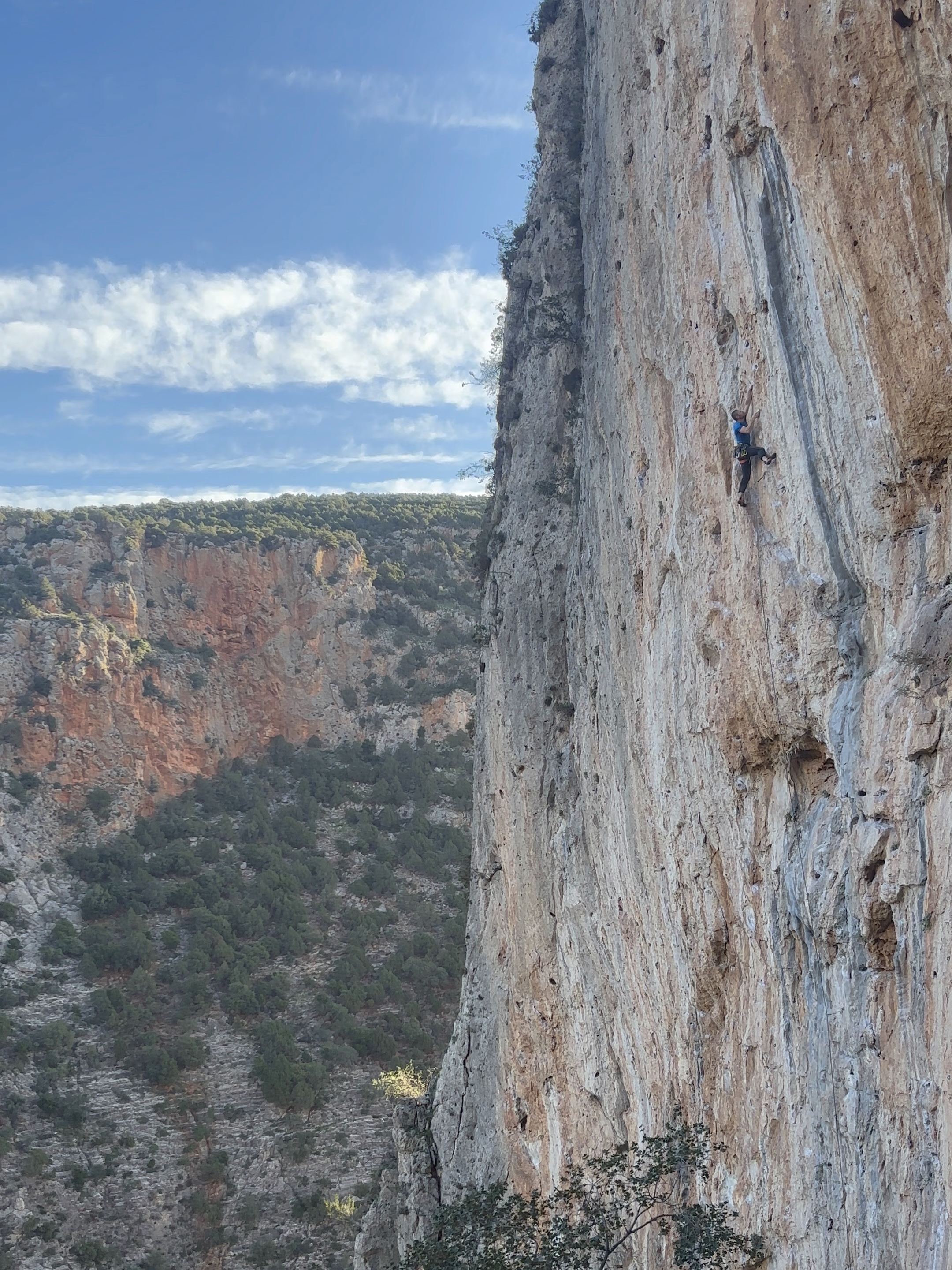 Climber in a blue t-shirt on a tall cliff, set within a valley with another crag on the opposite side.