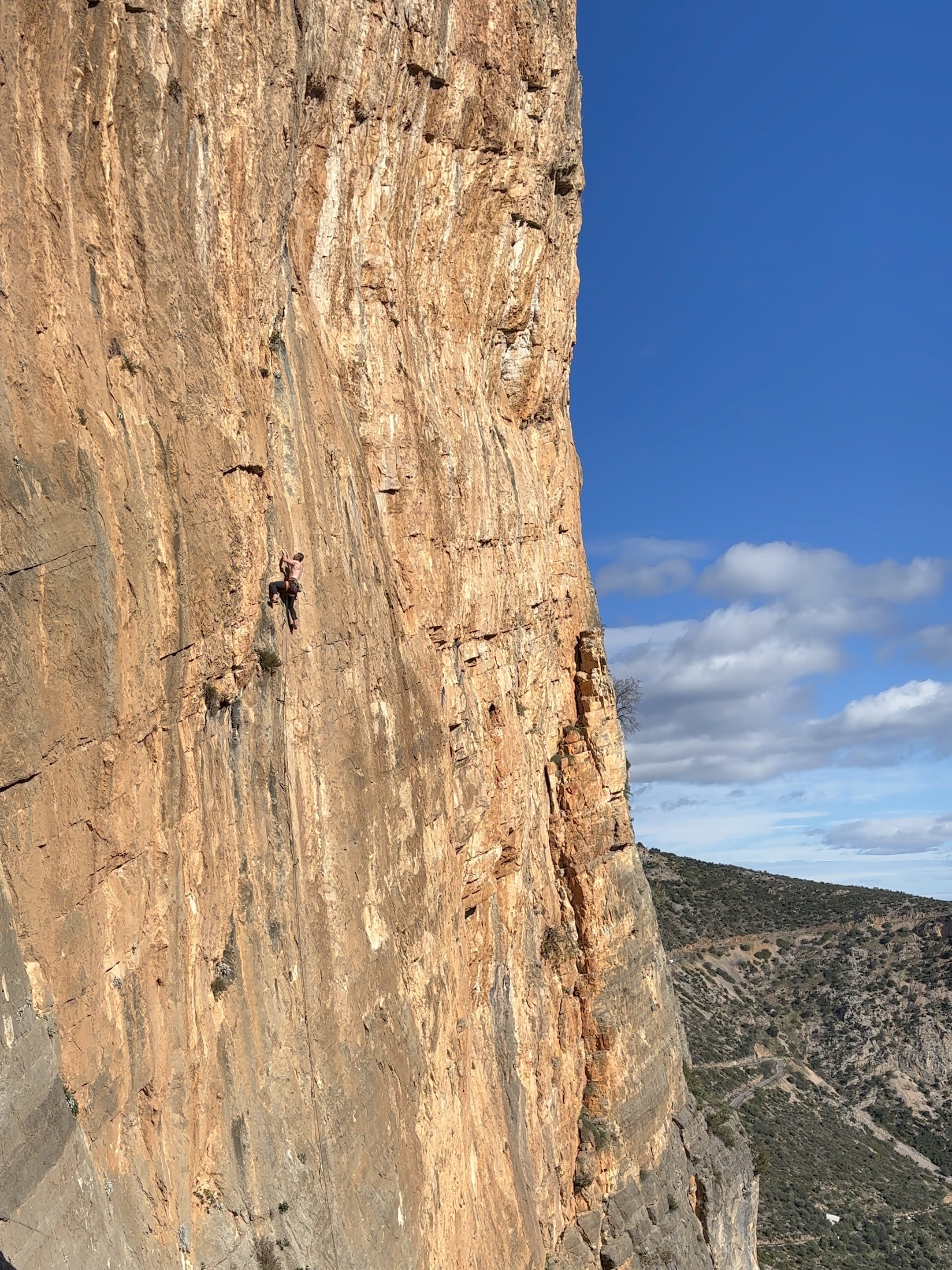 Climber using small crimpy holds on a vertical orange wall with a winding road up the mountain side in the background.