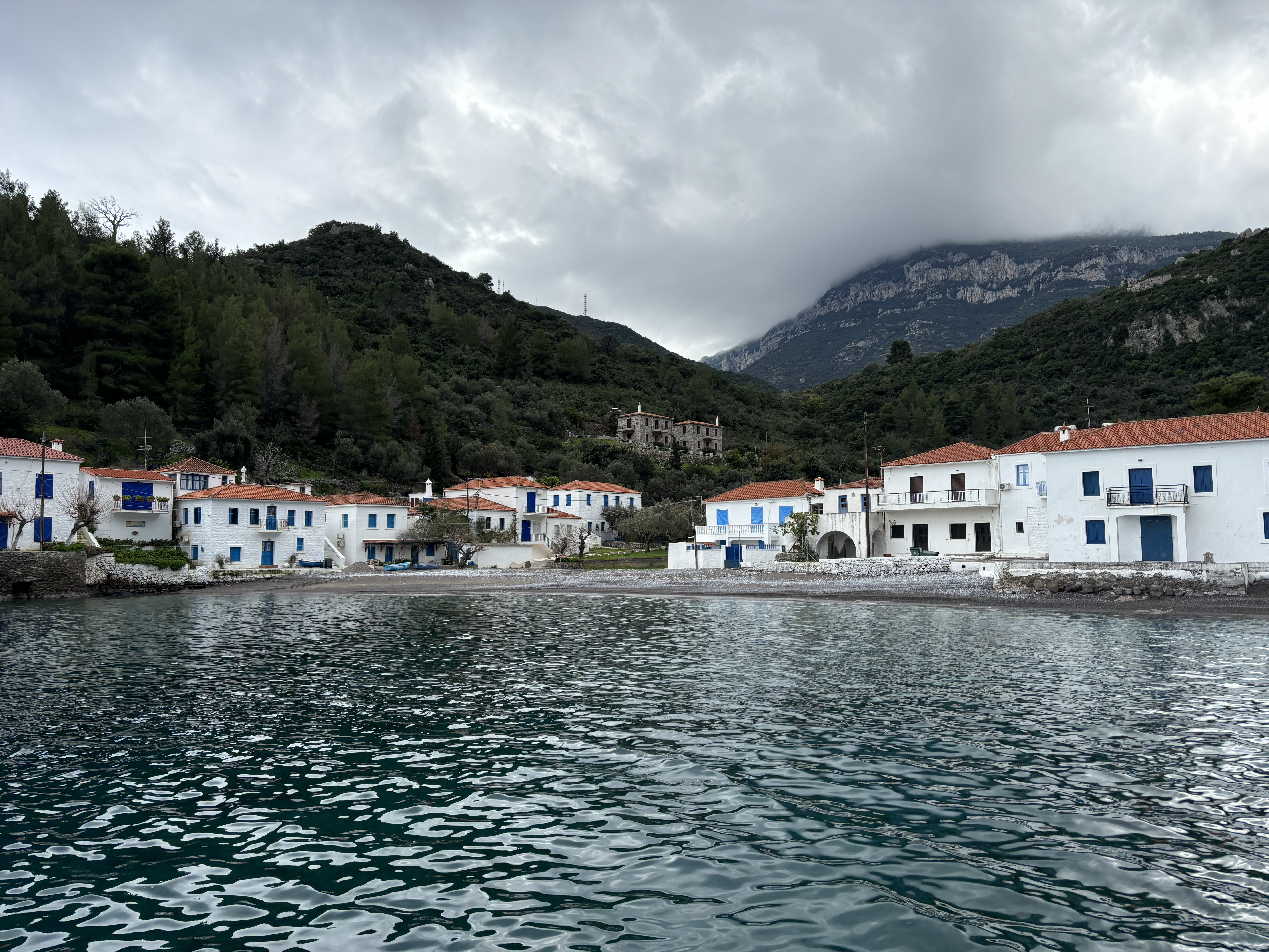 White washed houses with terracotta tiled roofs and blue shutters on the windows set on the sea front with mountains and limestone cliffs behind.