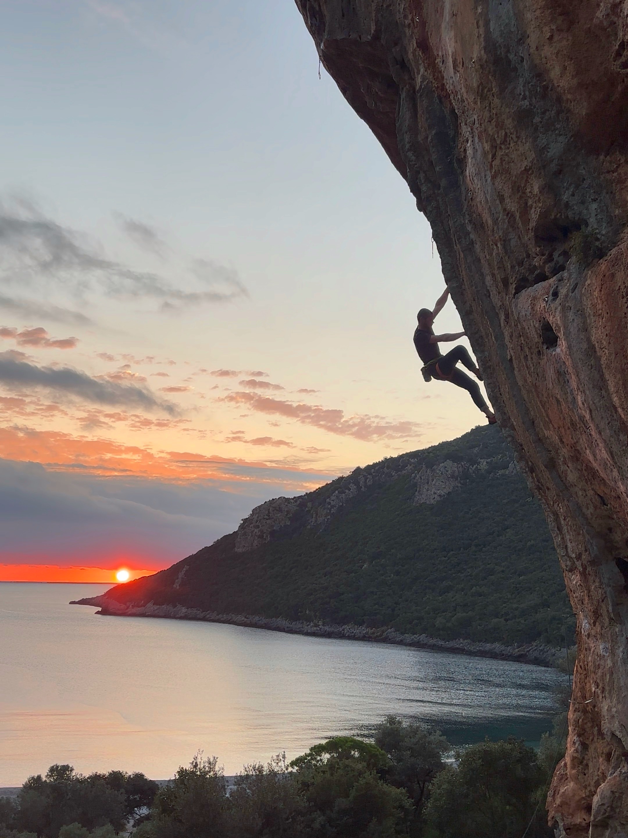 Climber silhouetted on a steep tufa route with the orange sun rising above a pebble bay and shimmering on the sea.