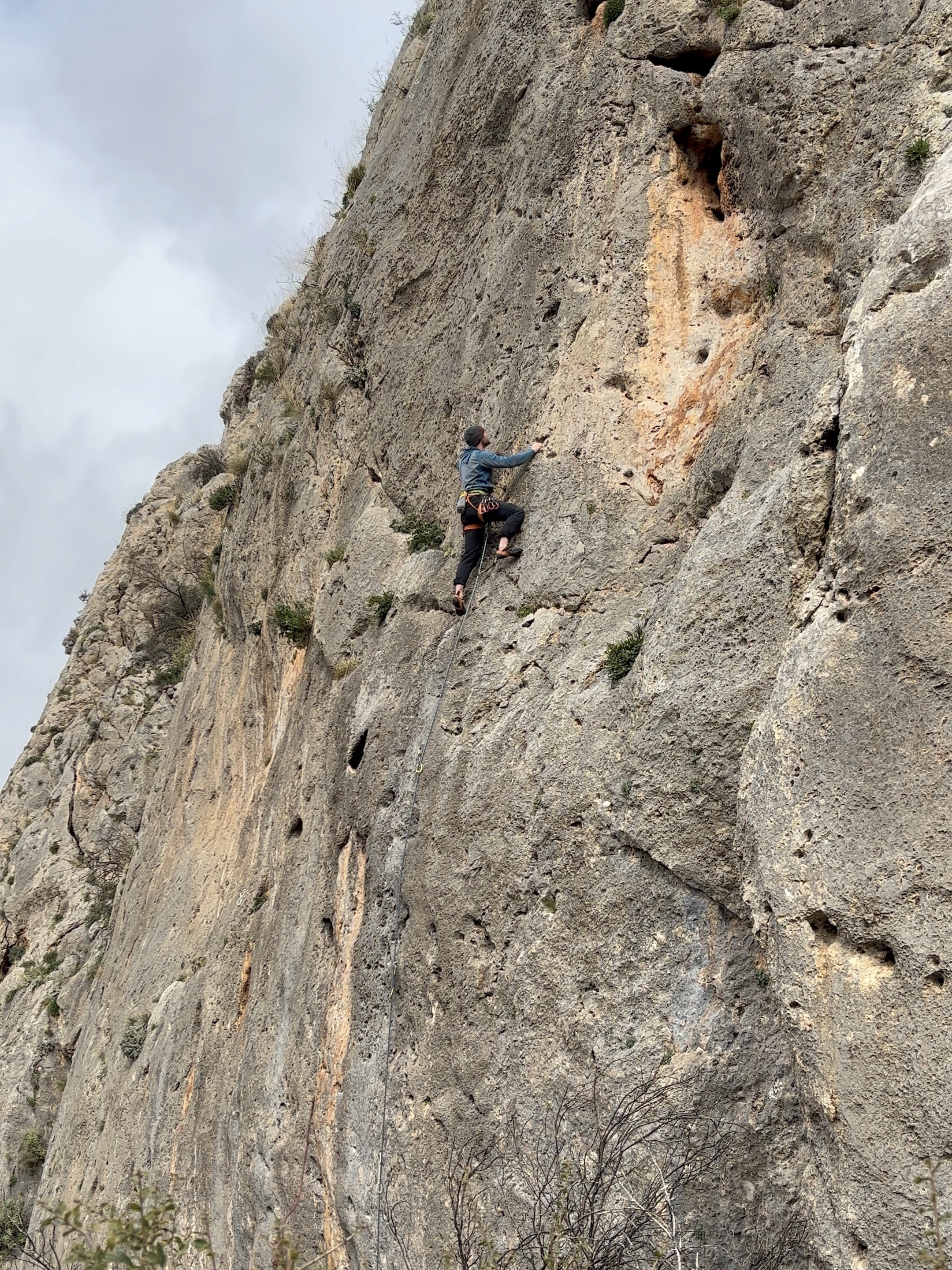Climber in grey hat and blue fleece on a grey slabby wall looking for the next hand hold on a cloudy day.