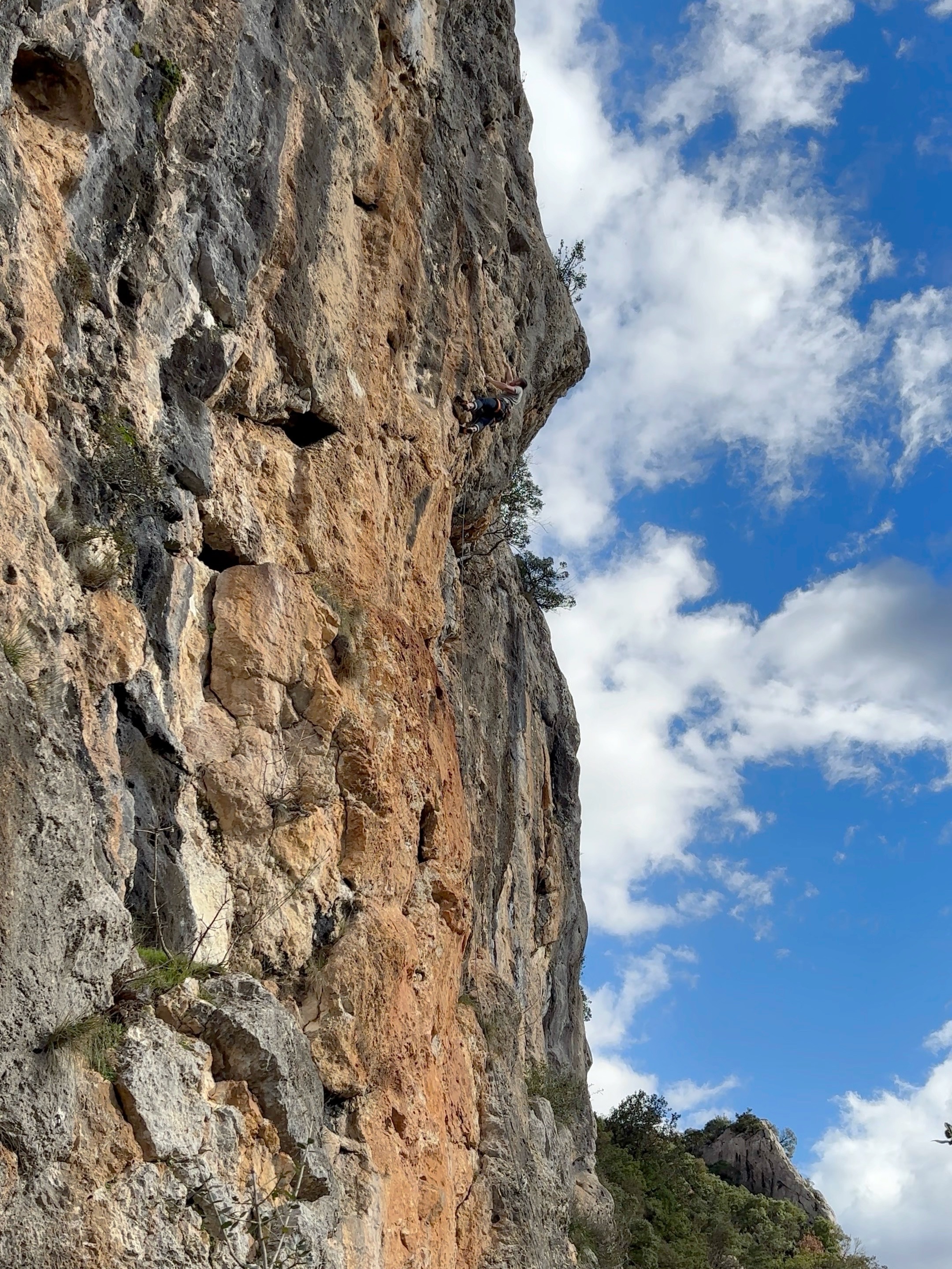 A climber performs the crux move in a near horizontal position on an orange and grey limestone cliff, with blue sky and cloud patches swirling in the background. 