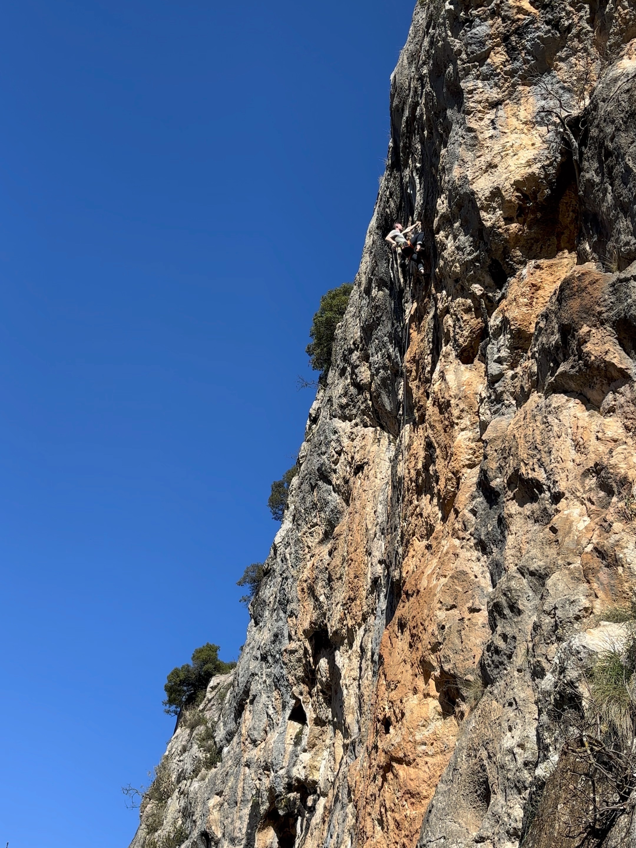 Climber on a steep grey and orange cliff, set against a vibrant blue sky.