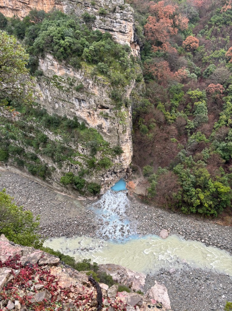 Vibrant blue thermal water meets the green/brown water of a river, as viewed from above. 
