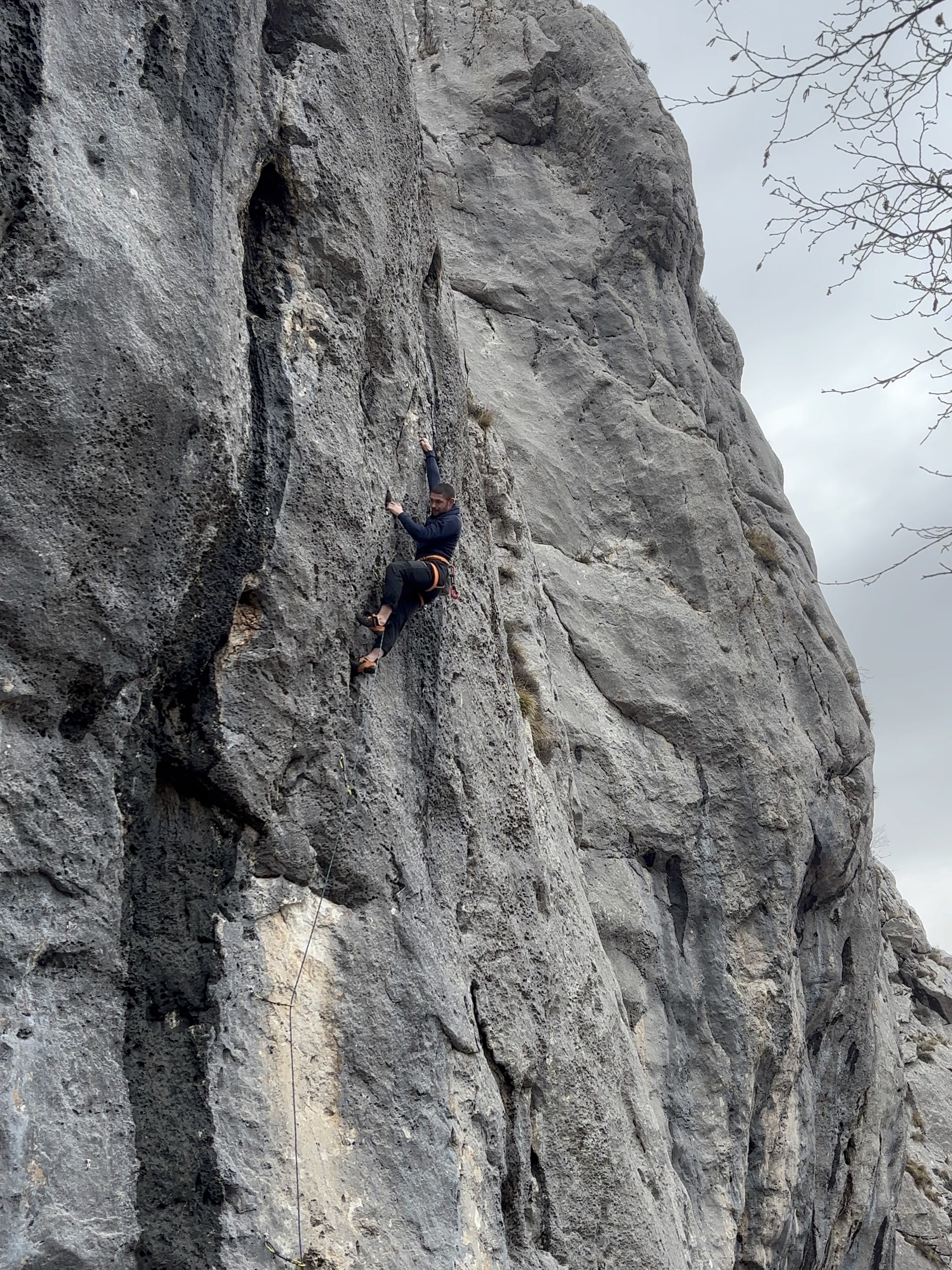 A climber performs a long reach on a gently overhanging grey limestone cliff. 