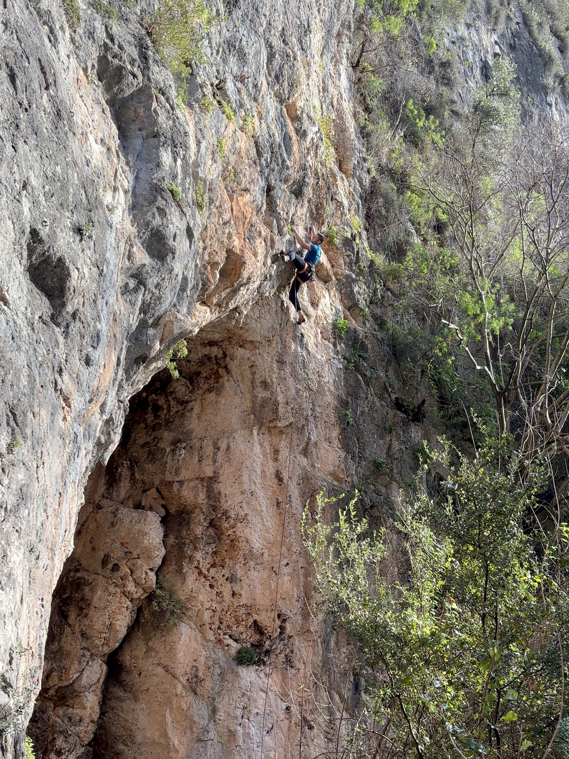 Climber in a blue shirt climbing a steep limestone wall, right of a cave. 