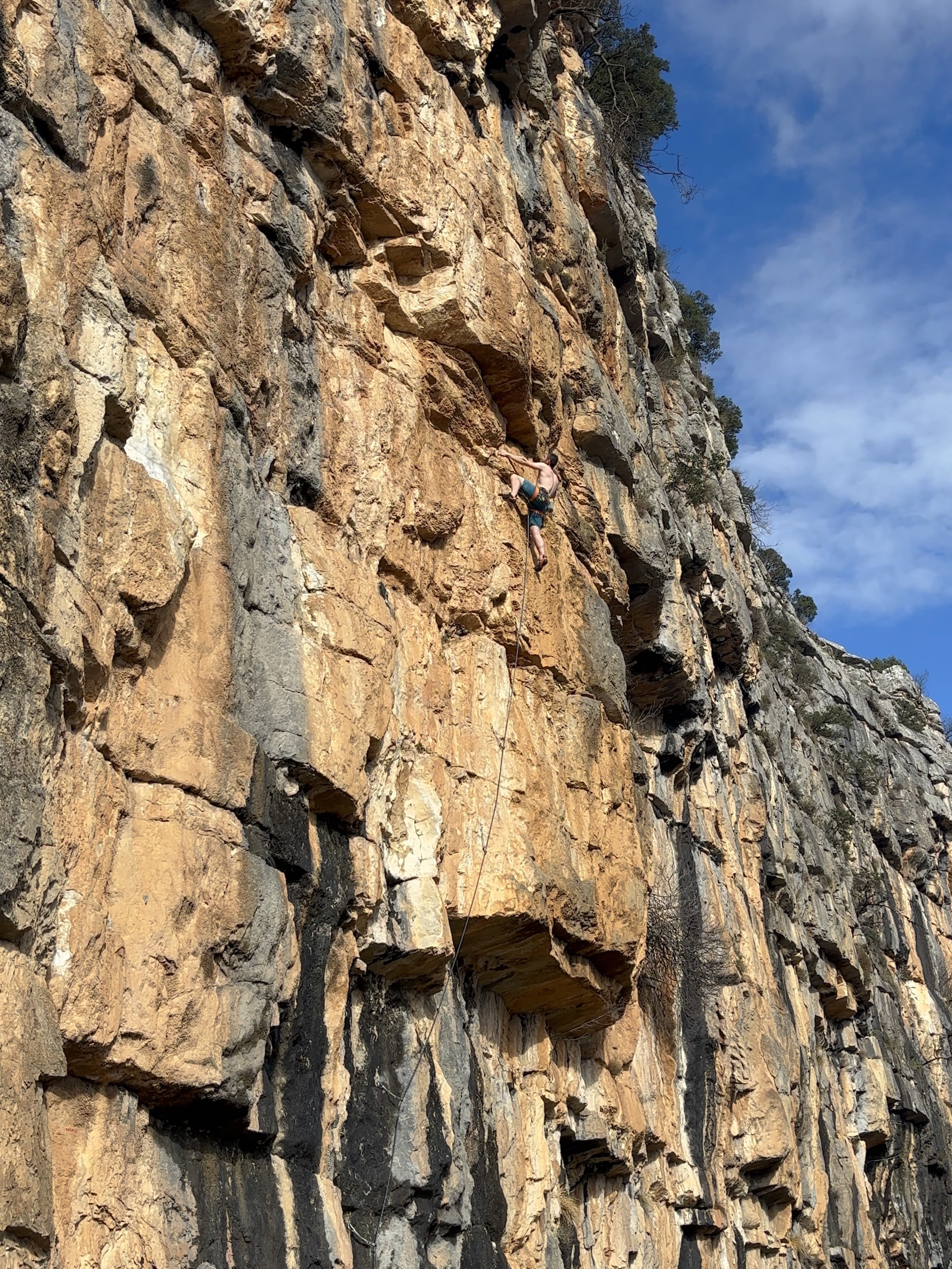 A shirtless climber eyes up the next moves on orange limestone cliff with black and grey streaks. 