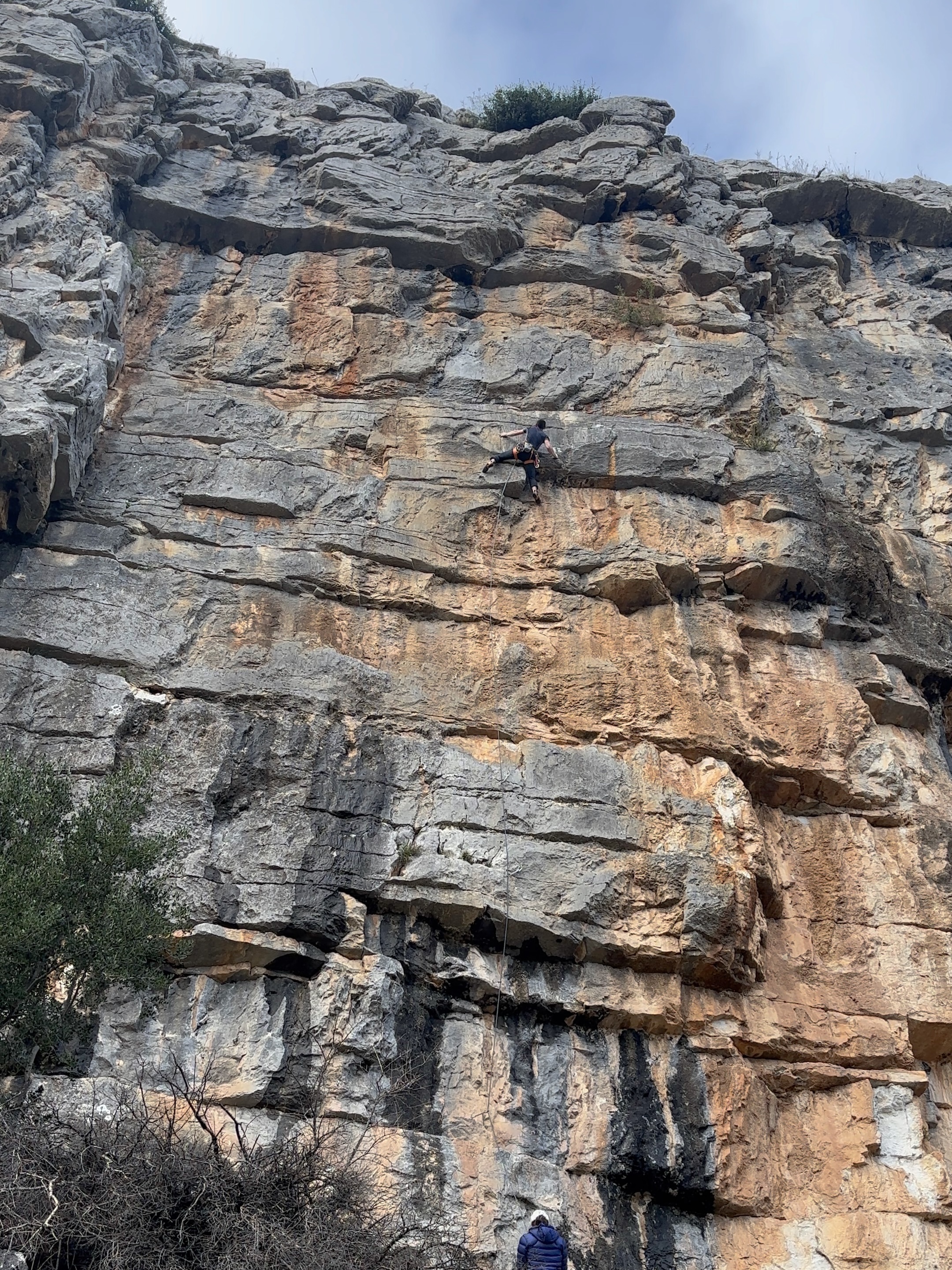 A front on view of a climber tackling the crux move on a 40 meter long cliff with several horizontal breaks interspersed by thin crimpy climbing. 