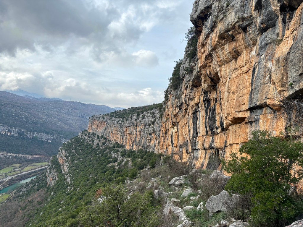 A long and continuous band of cliffs with hills in the background and a river and road in the canyon below. 