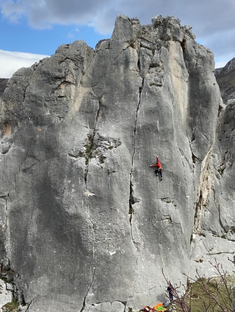 A climber in red balances on tiny holds on a technical grey limestone slab, on a large fin of rock. 