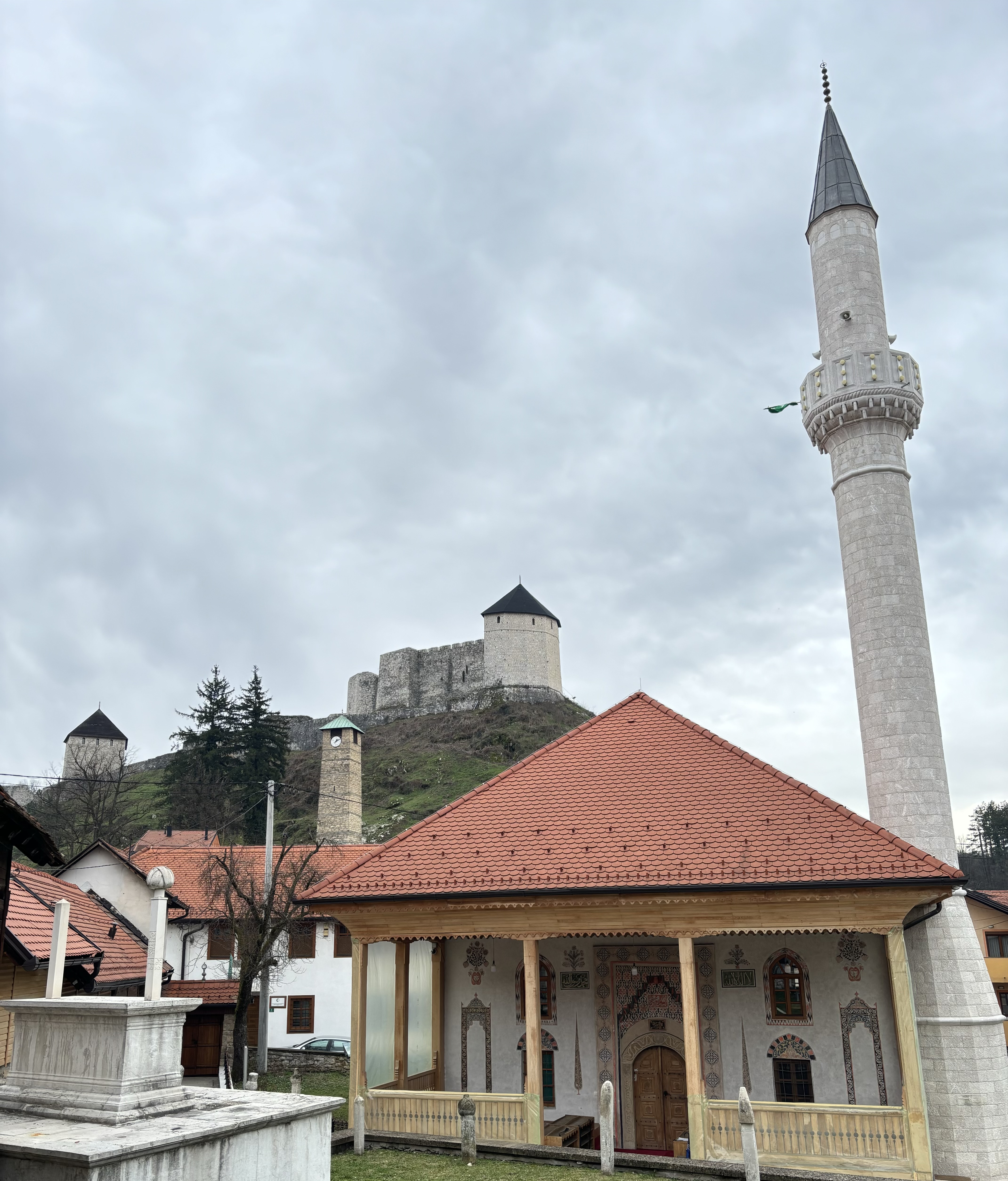 A beautifully decorated historical mosque and minaret sits below an imposing hilltop castle fortress with a clock tower between the two. 
