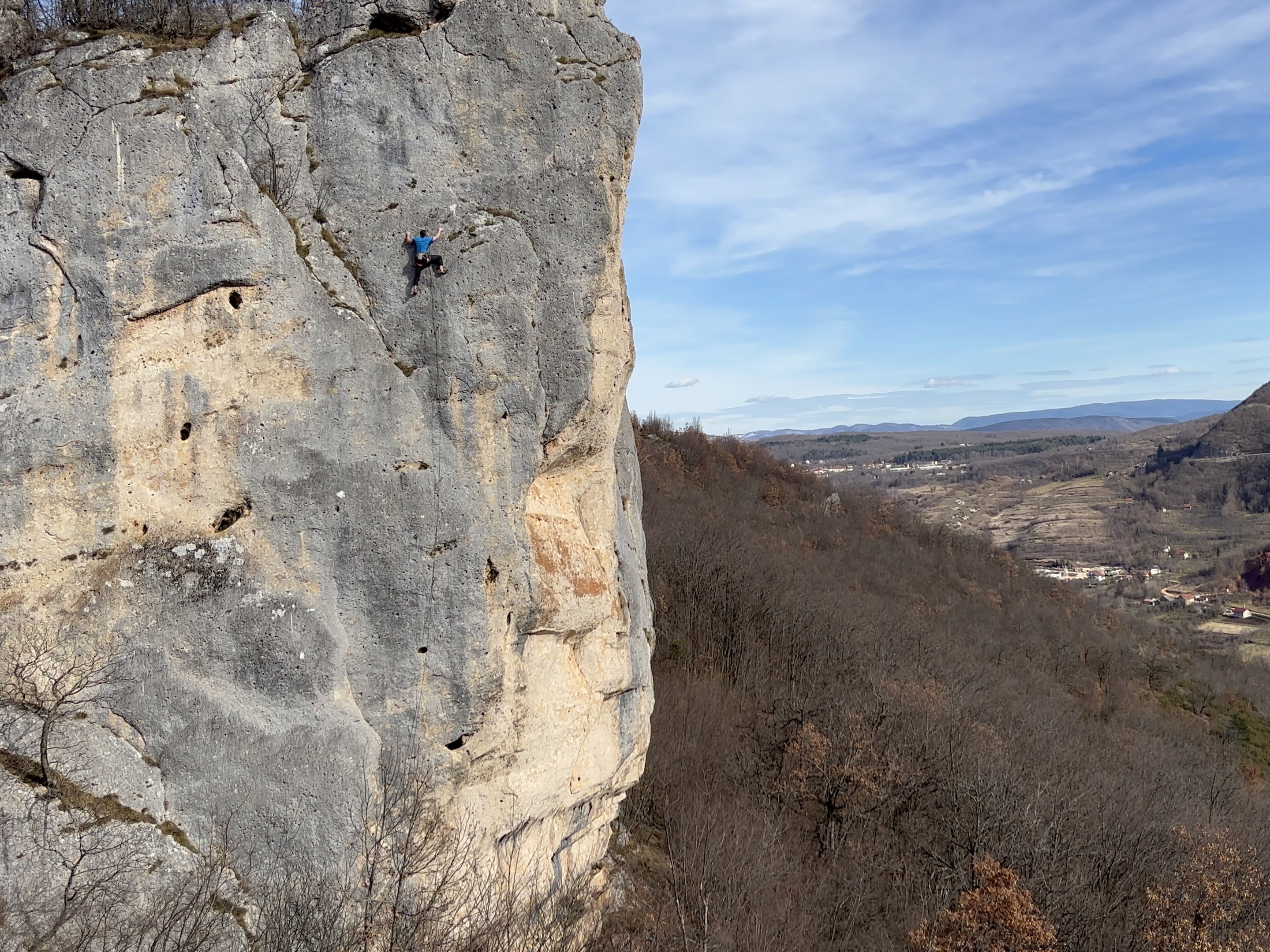 A climber in a blue t-shirt on a large conglomerate limestone cliff that projects out of the hillside, with a forest below and a green valley in the background. 
