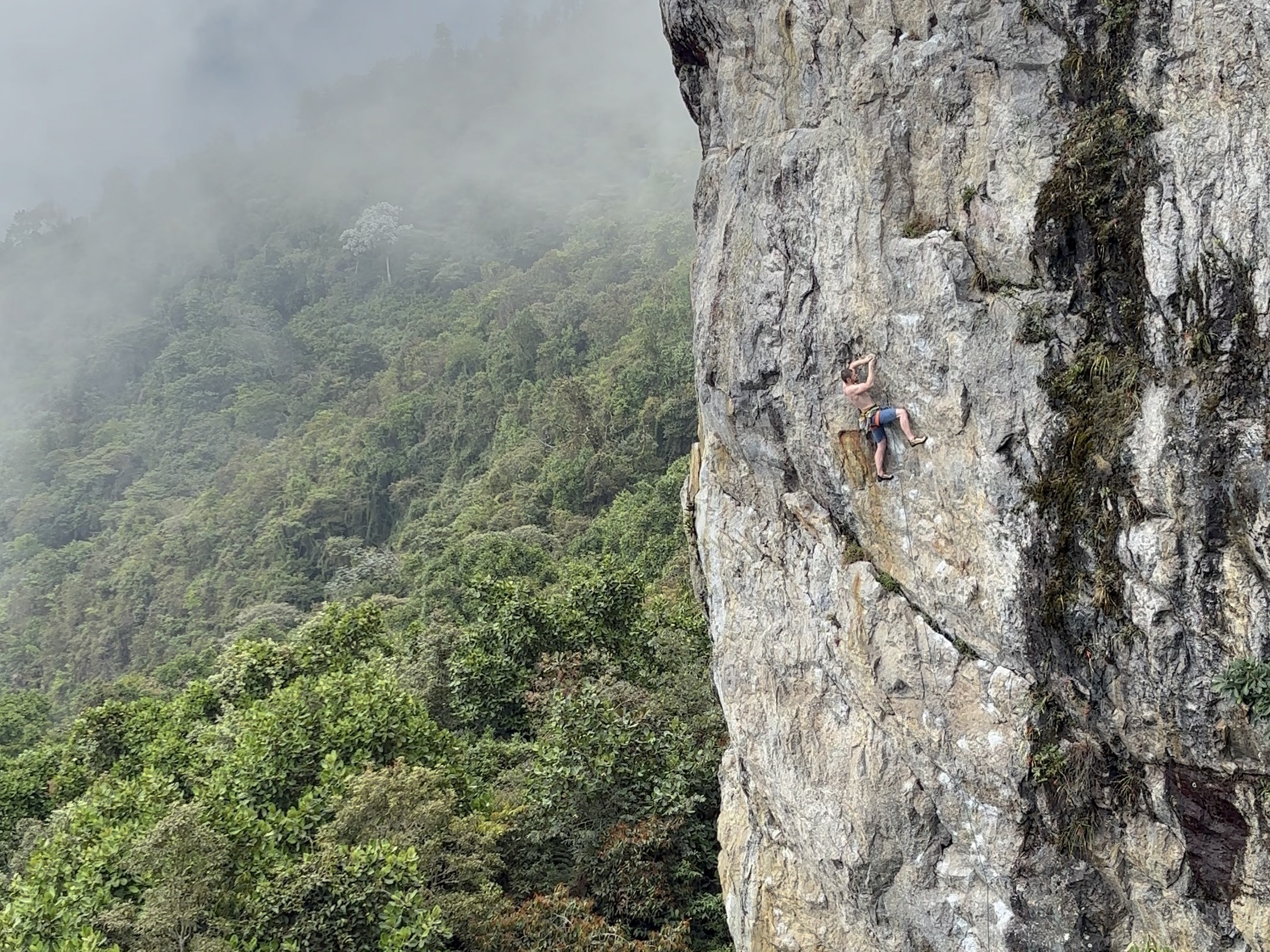 A climber on a steep granite cliff above the misty jungle below. 